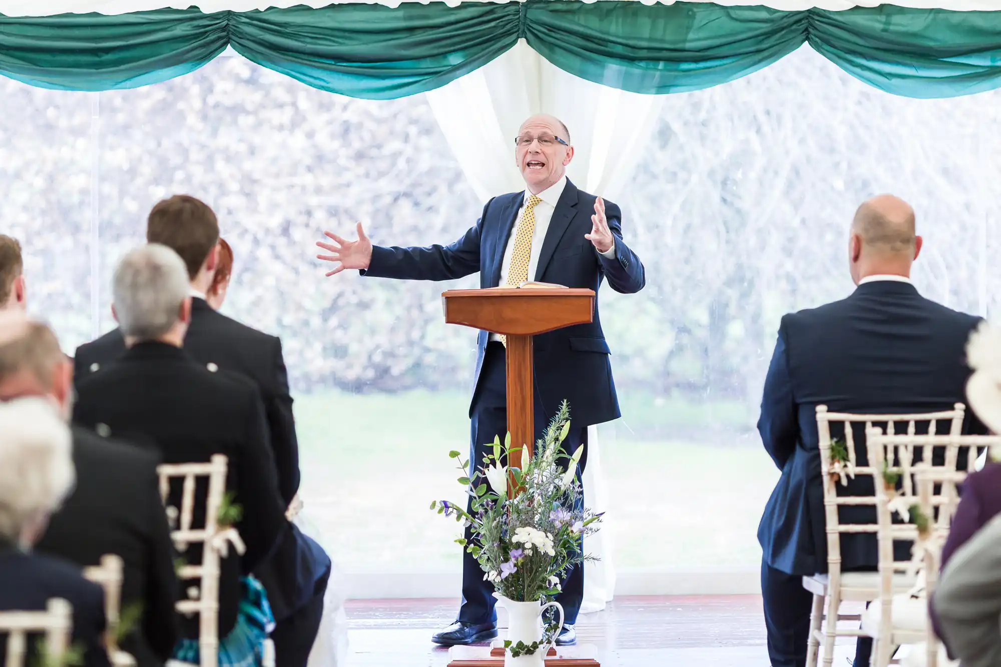 A man in a suit and yellow tie stands at a lectern, gesturing energetically whilst speaking to a seated audience at an indoor event. Green curtains and a flower arrangement decorate the bright, windowed space.