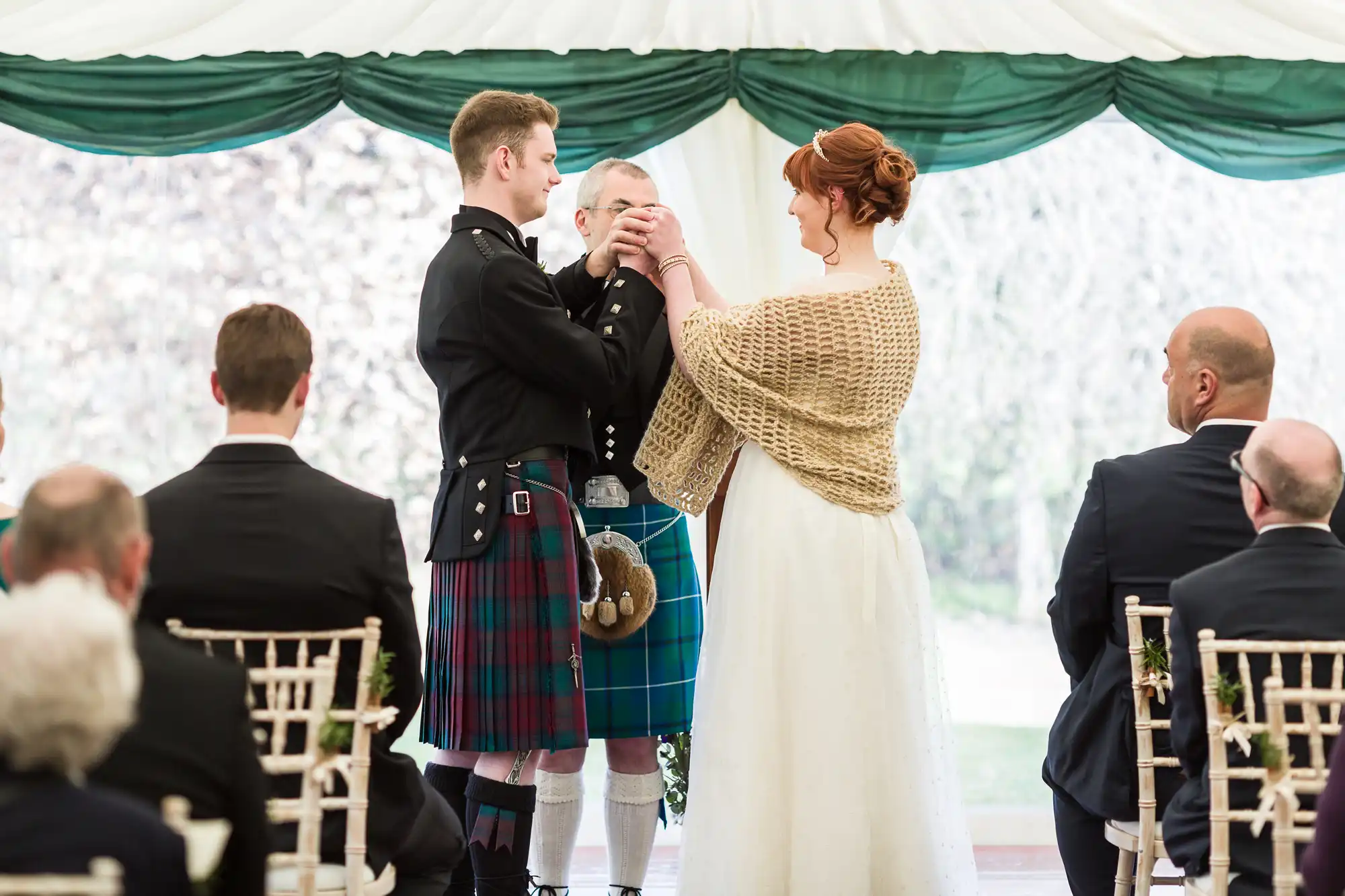 A bride and groom stand holding hands during a wedding ceremony. The groom wears a kilt and sporran, the bride wears a white dress with a tan shawl. A man in a tartan kilt stands behind them. Seated guests watch in a white marquee with green drapes.