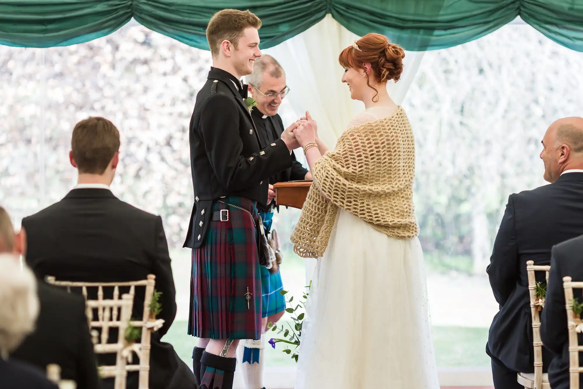 A smiling couple exchange rings at a wedding ceremony under a green and white marquee. The groom wears a traditional Scottish kilt and jacket, while the bride wears a white dress with a tan shawl. Guests in formal attire are seated, and an officiant stands between them.