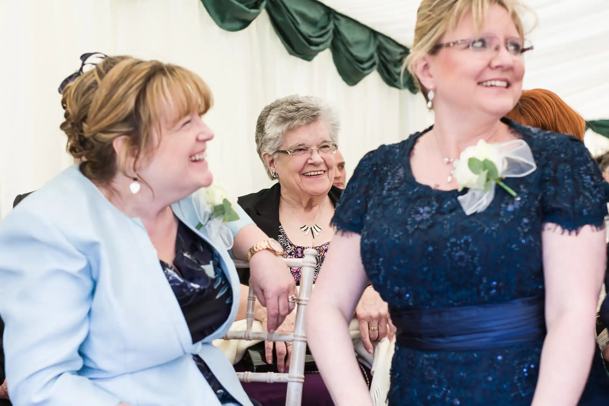 Three older women smiling and laughing together at an indoor event, possibly a wedding. Two women in the foreground wear formal dresses with corsages, while a third woman behind them, in glasses and a patterned top, also smiles warmly.