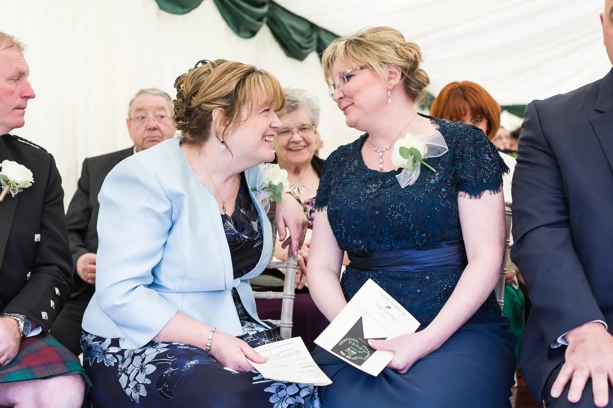 Two women dressed formally sit closely together, smiling and talking, holding programmes and flowers. They are surrounded by other seated guests at an indoor event, likely a wedding, with white and green draped fabric in the background.