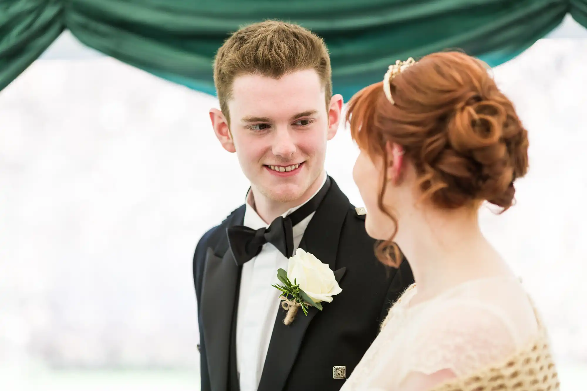 A young man in a tuxedo with a white rose buttonhole smiles warmly at a woman with red hair in an updo, wearing a delicate hairpiece and light-coloured dress. They stand together under green drapery, suggesting a formal or wedding setting.