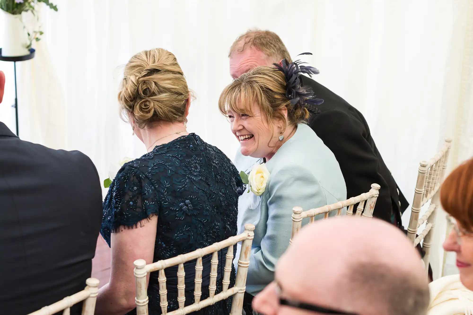 A woman in a light blue jacket with a white rose corsage and feathered fascinator smiles whilst seated on a white chair at an indoor event. She is surrounded by others in formal attire, and the background features white drapes.