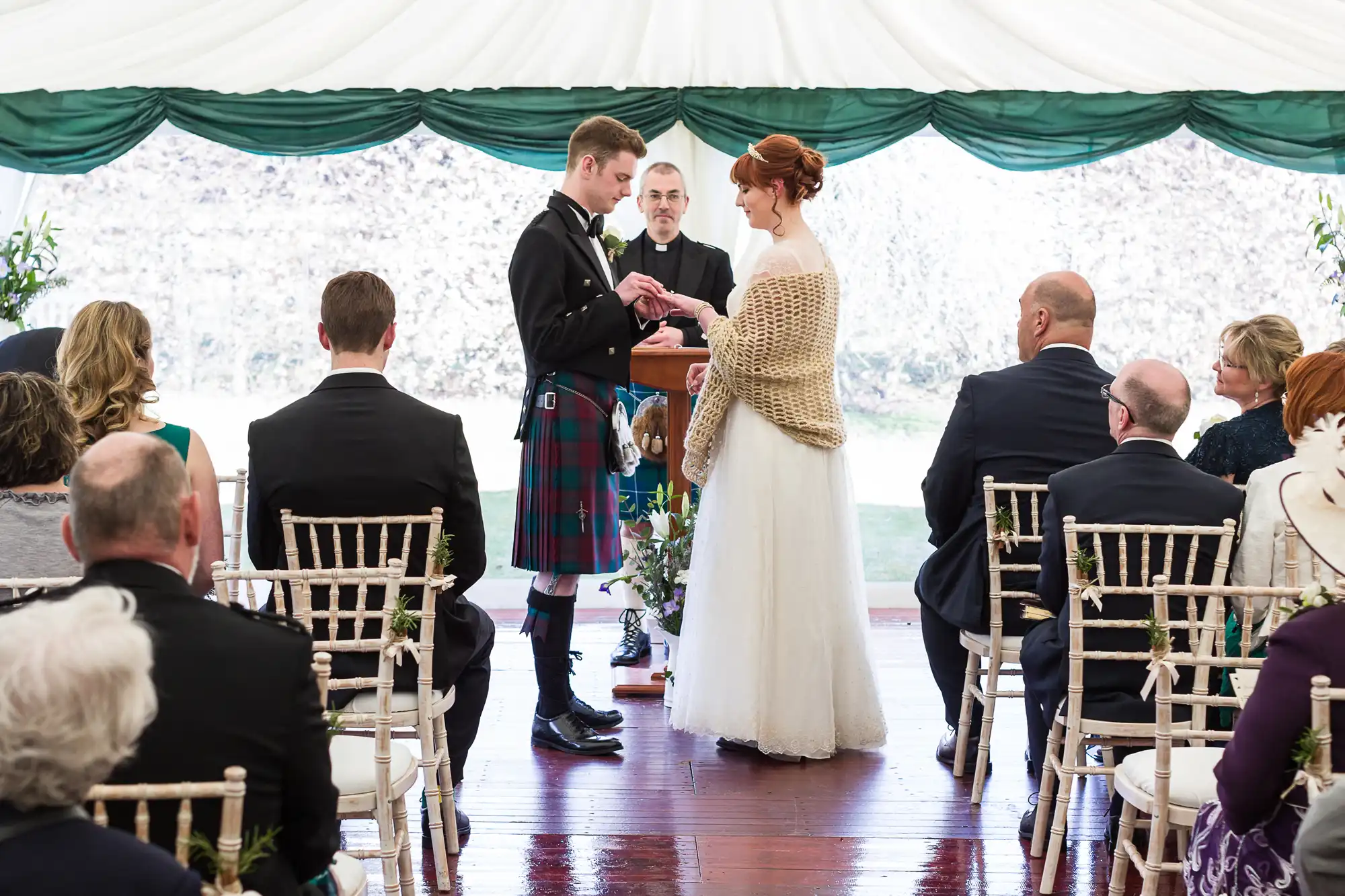 A bride and groom stand facing each other in a white marquee, exchanging rings during their wedding ceremony. The groom wears a kilt; the bride wears a white dress and shawl. Guests sit on white chairs, and an officiant stands behind the couple.