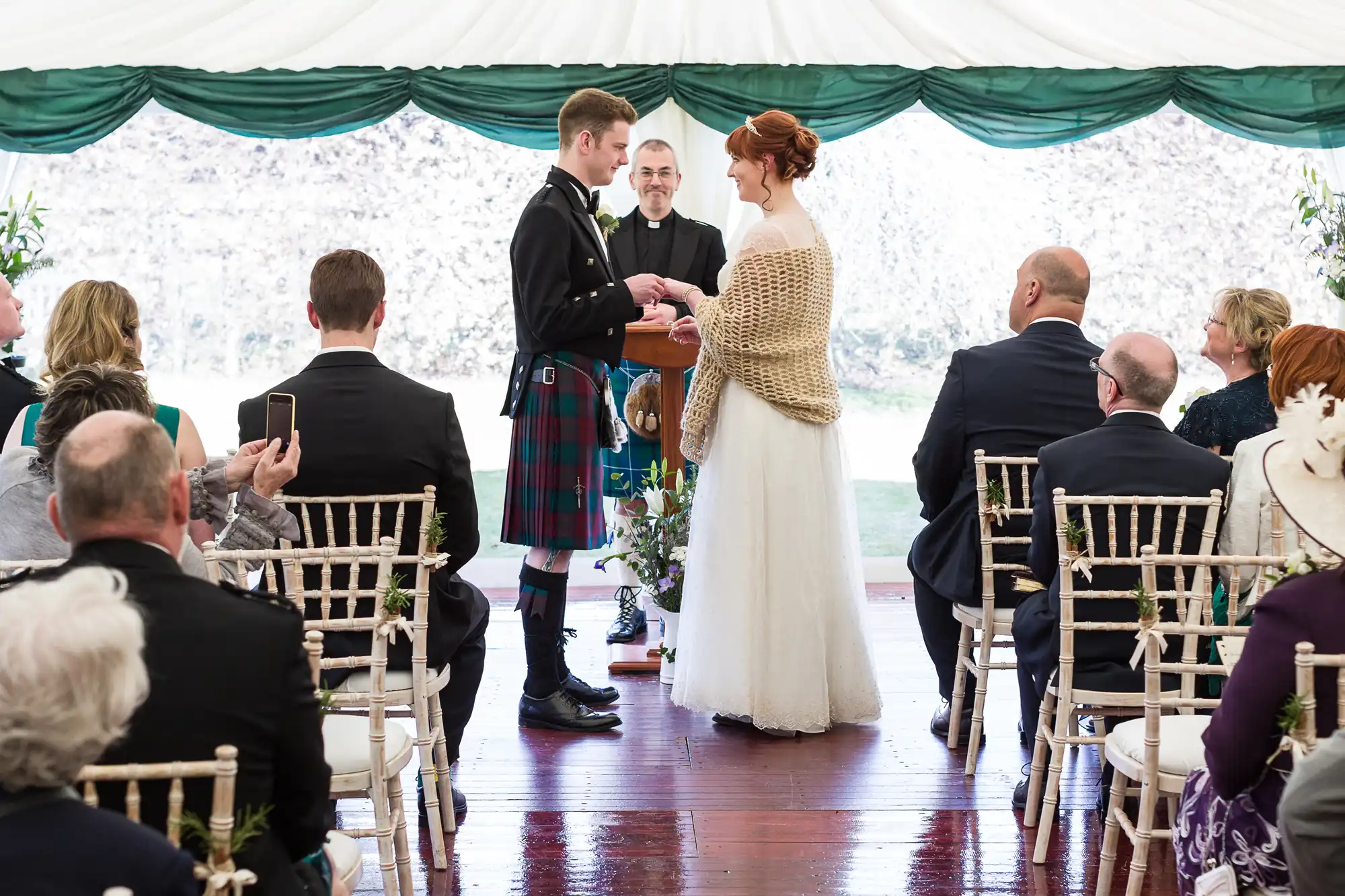 A bride and groom stand facing each other under a white marquee during their wedding ceremony. The groom wears a kilt, and the bride wears a white dress with a shawl. Guests sit on white chairs, watching, whilst an officiant stands behind the couple.