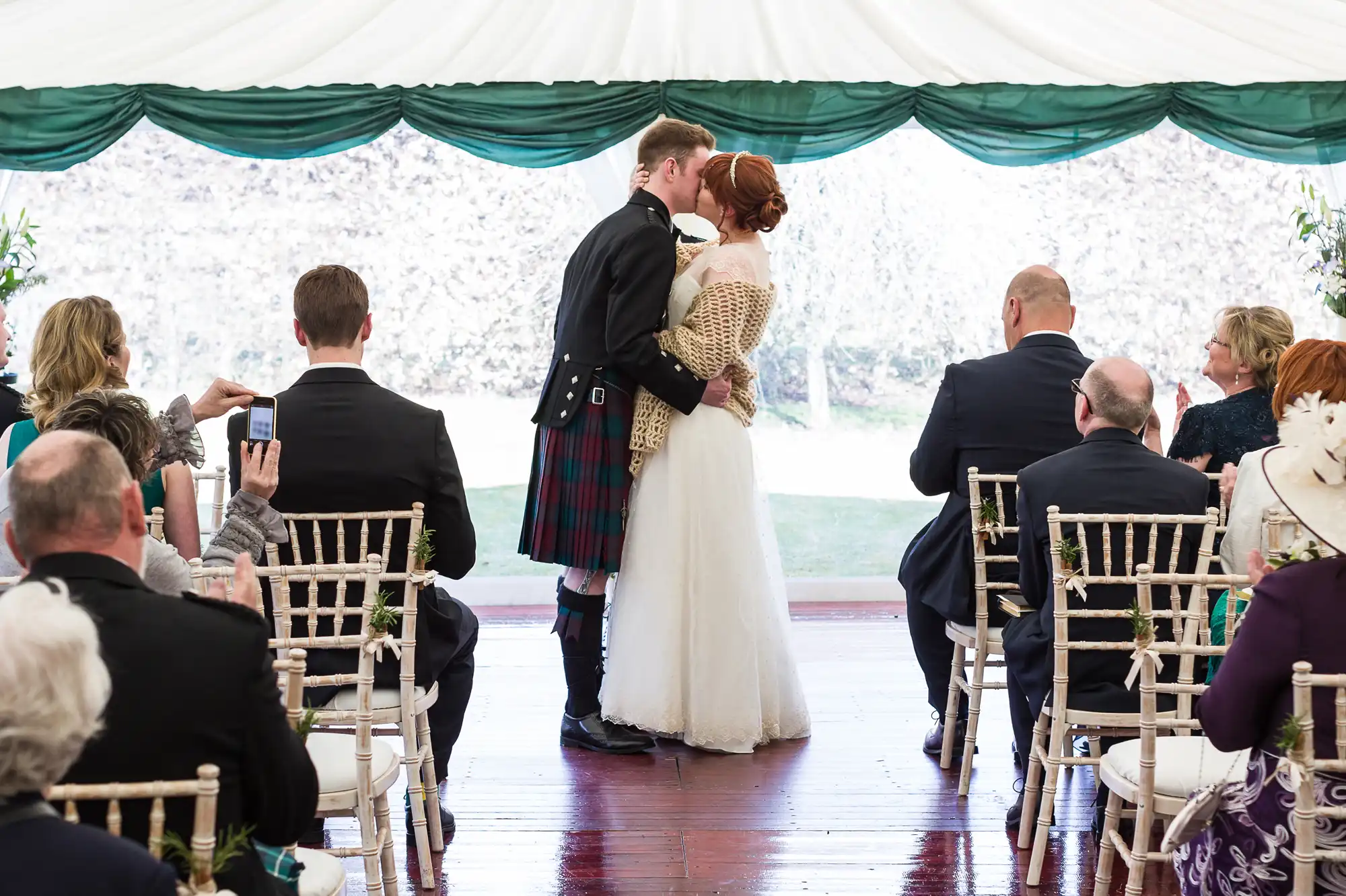A bride and groom share a kiss at the altar during their wedding ceremony in a marquee. The groom wears a black jacket and tartan kilt; the bride wears a white dress and shawl. Guests are seated, facing them, some taking photos with their mobiles.