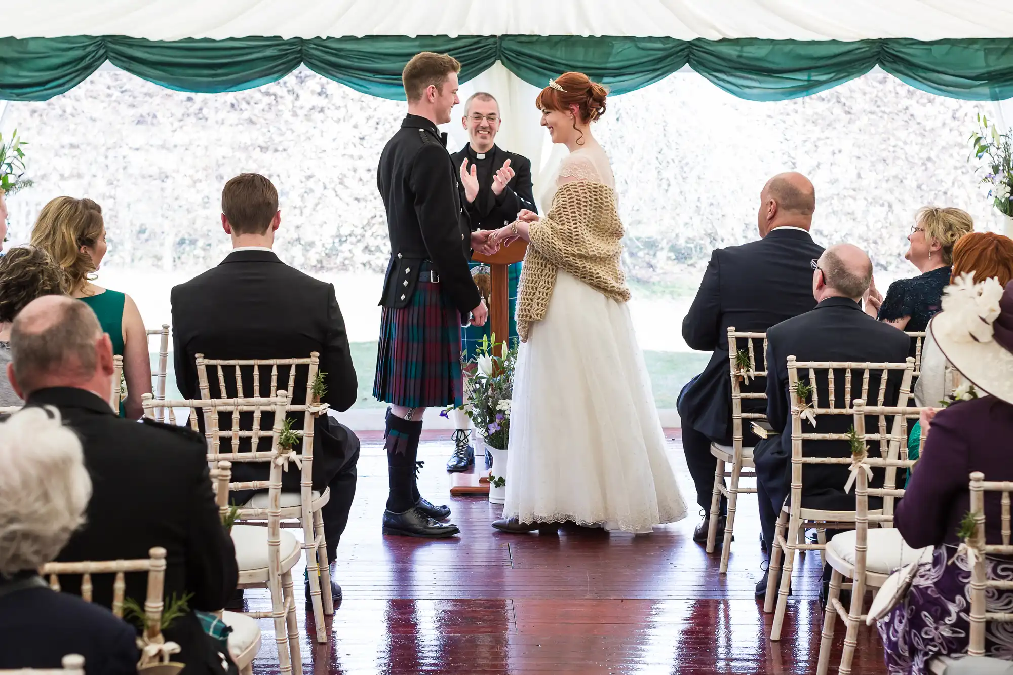 A bride and groom stand facing each other under a white marquee during their wedding ceremony. The groom wears a kilt, and the bride wears a white gown with a shawl. Guests sit on light chairs, and an officiant stands smiling behind the couple.