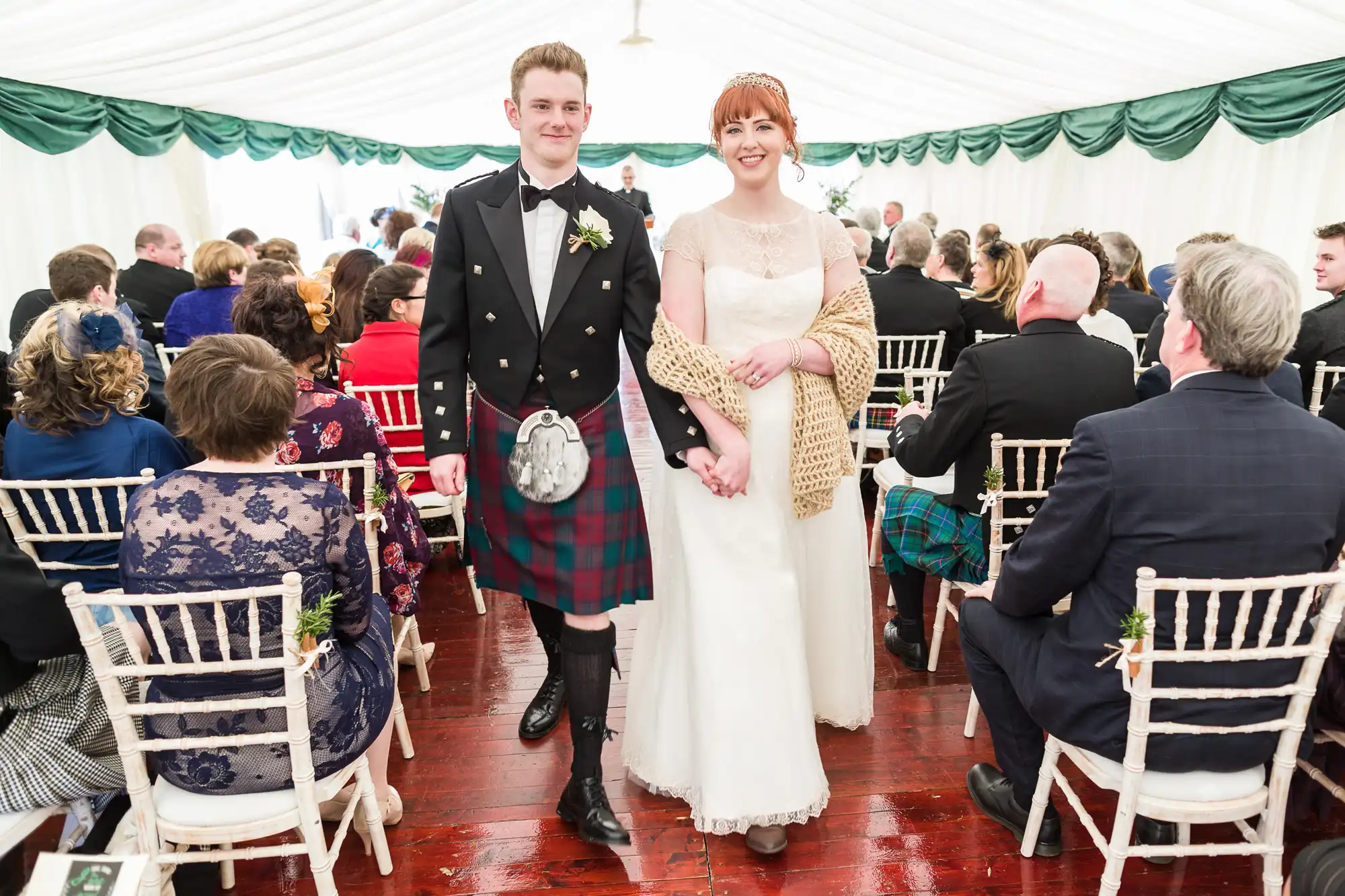 A smiling bride in a white dress and a groom in a Scottish kilt walk arm in arm down the aisle inside a white marquee, surrounded by seated wedding guests dressed formally, some in kilts. The floor is wooden and the chairs are white.