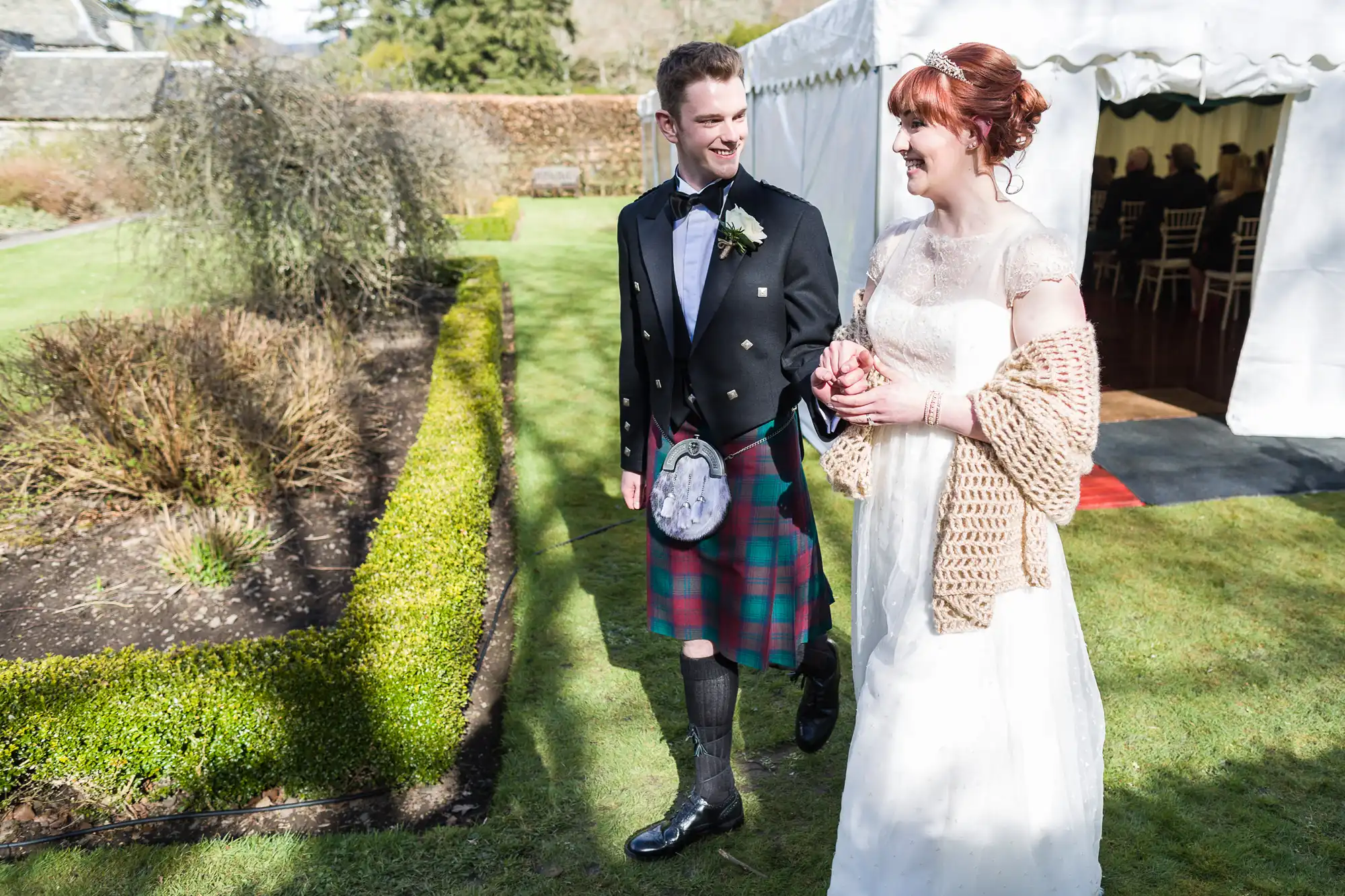 A smiling bride in a white dress and shawl walks hand-in-hand with a groom in a black jacket and tartan kilt outside near a garden bed and white marquee, both looking at each other on a sunny day.