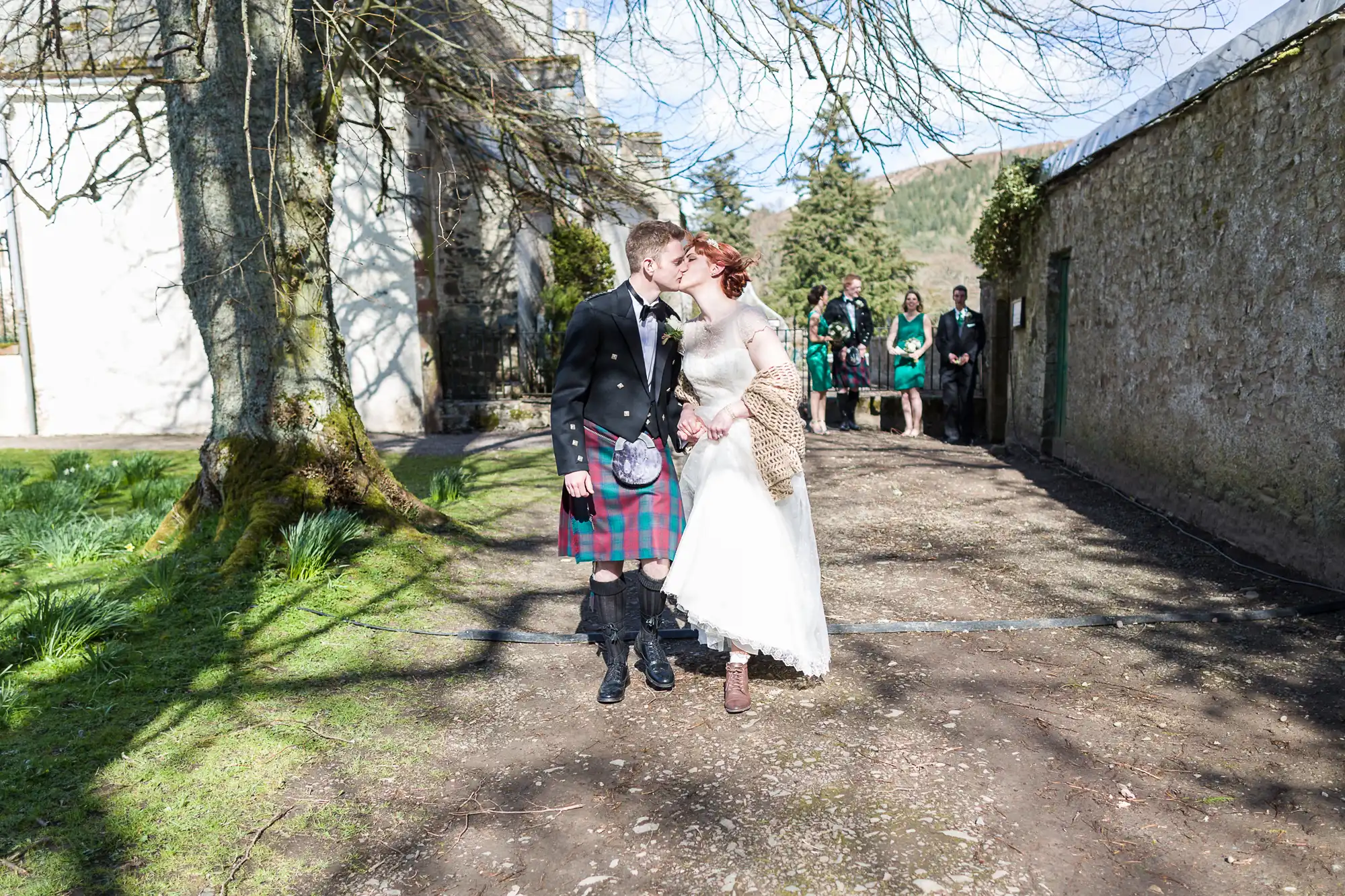 A couple in wedding attire share a kiss outdoors; the groom wears a kilt and jacket, the bride wears a long white dress and brown shawl. Sunlight filters through trees. Wedding guests stand in the background near a stone building and wall.