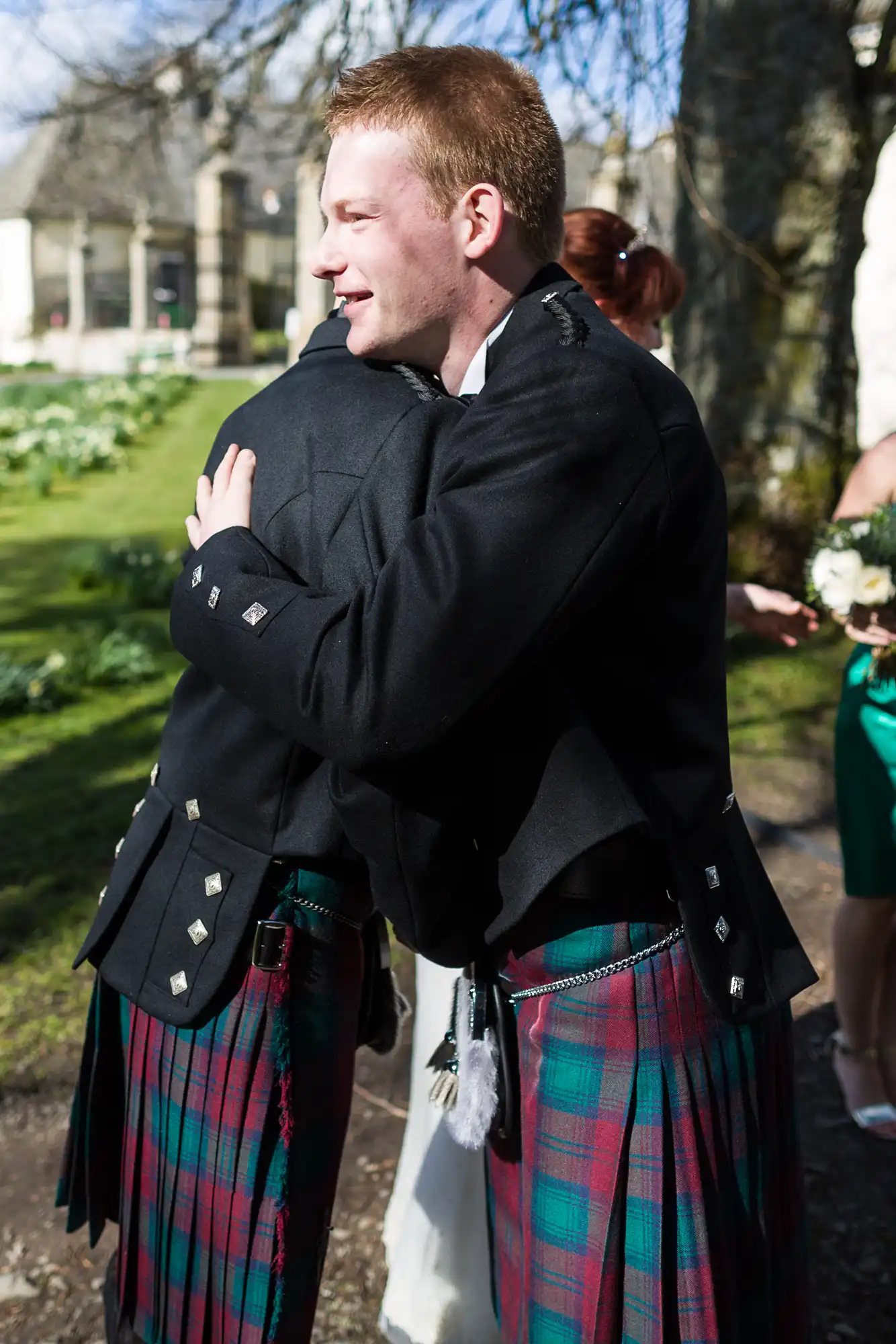 Two men wearing traditional Scottish kilts and black jackets embrace outdoors on a sunny day. One man faces the camera smiling, whilst the other’s back is turned. Trees, green grass, and white flowers are visible in the background.