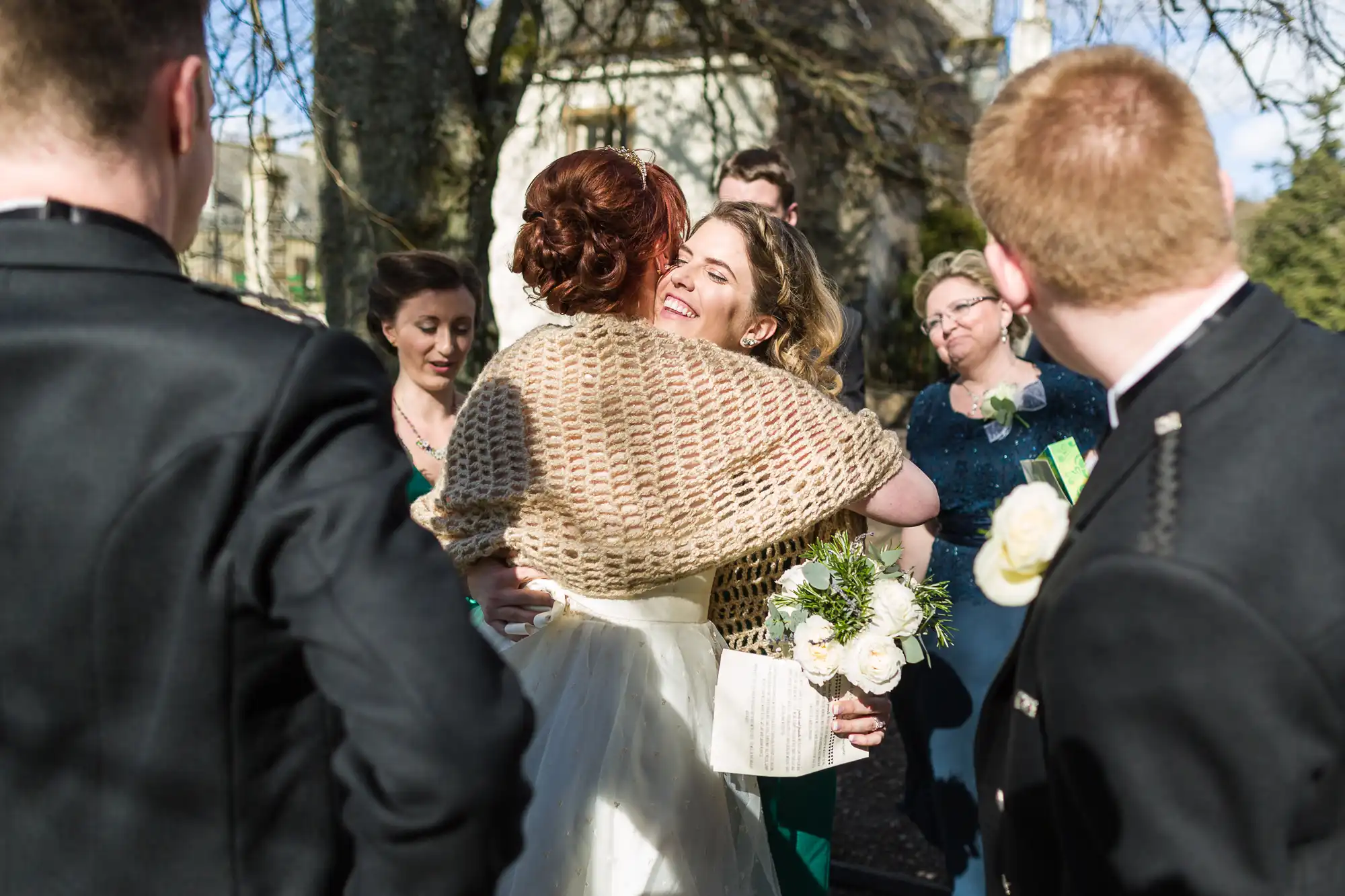 A bride in a white dress and tan shawl hugs a smiling woman in a gold shawl, holding a bouquet. Several people in formal attire, including men in kilts, stand around them outdoors under sunlight and trees, looking on happily.