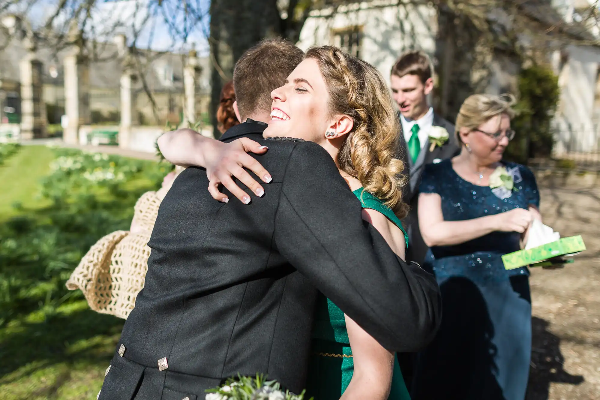 A woman in a green dress smiles warmly as she hugs a man in a dark suit outdoors. Other people in formal attire stand nearby under sunlight, surrounded by greenery and trees, suggesting a celebratory event like a wedding.