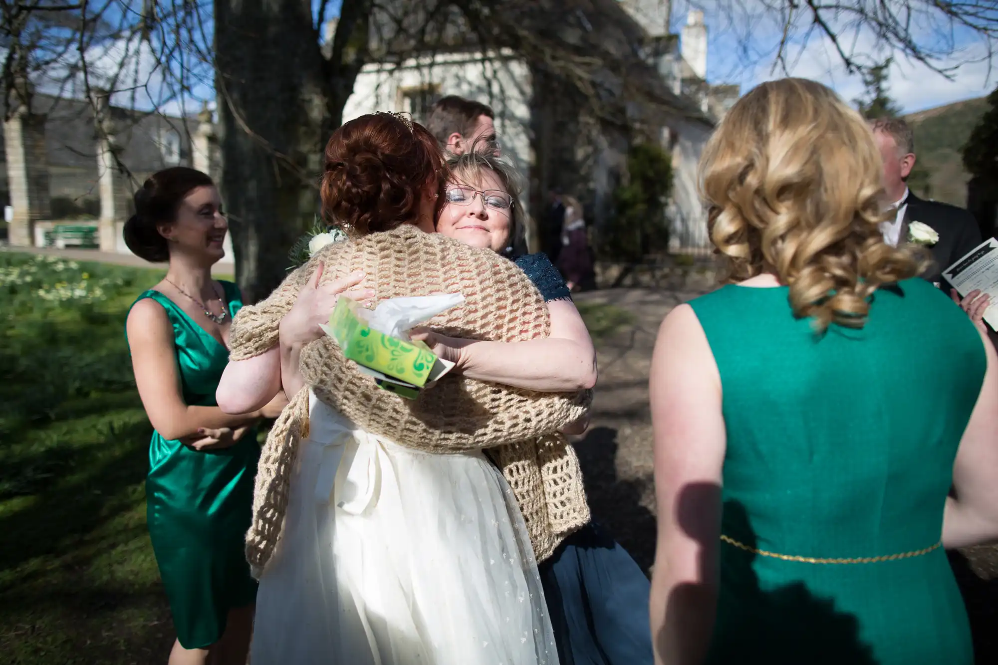 A bride in a white dress hugs an older woman wearing glasses and a beige crocheted shawl outdoors. They are surrounded by three women in green dresses, all smiling, with sunlight filtering through trees and a large building in the background.