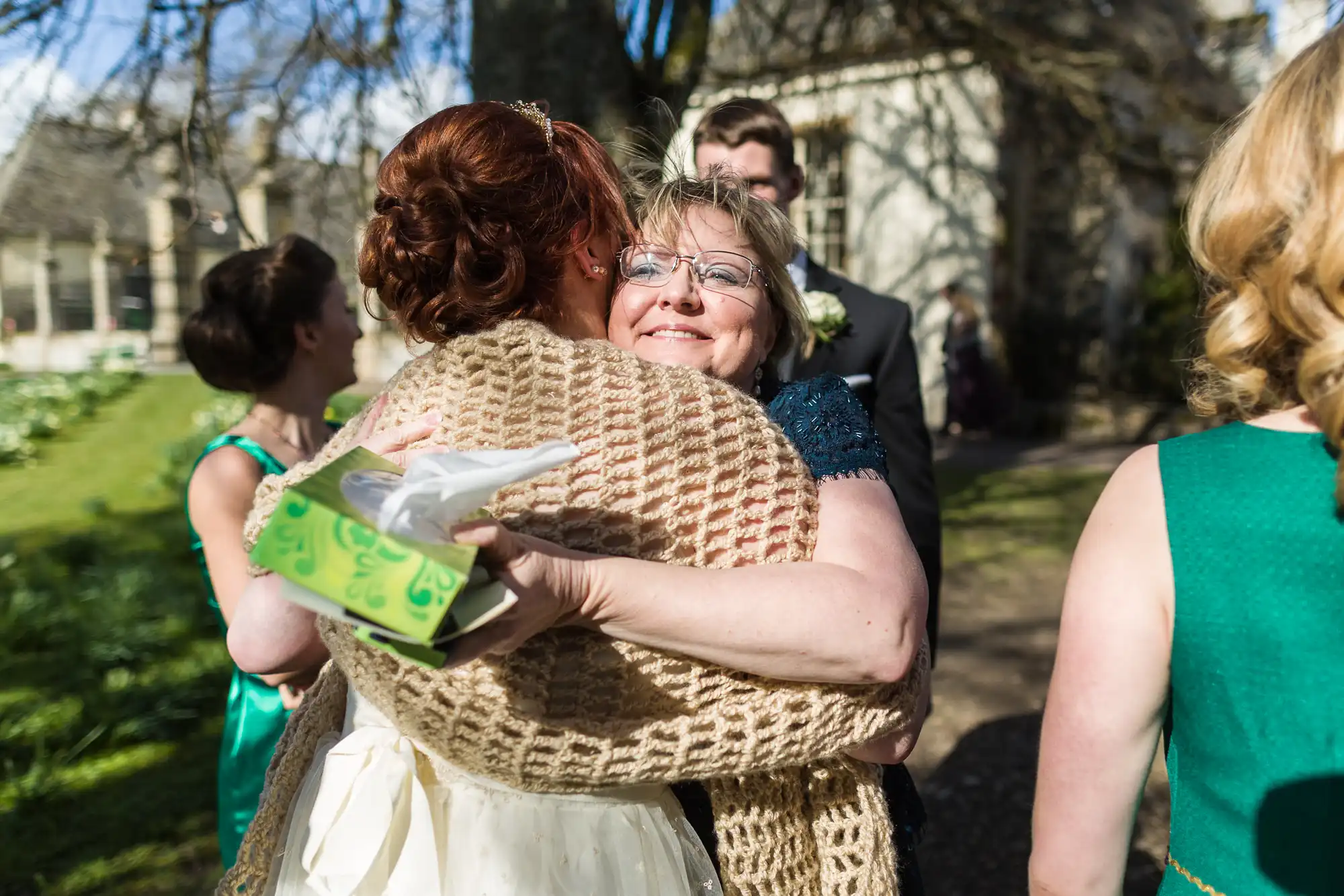 A woman in glasses hugs a bride outdoors on a sunny day, both smiling warmly. The bride holds a green tissue box and wears a cream shawl. Other people in formal attire stand in the background among trees and sunlight.