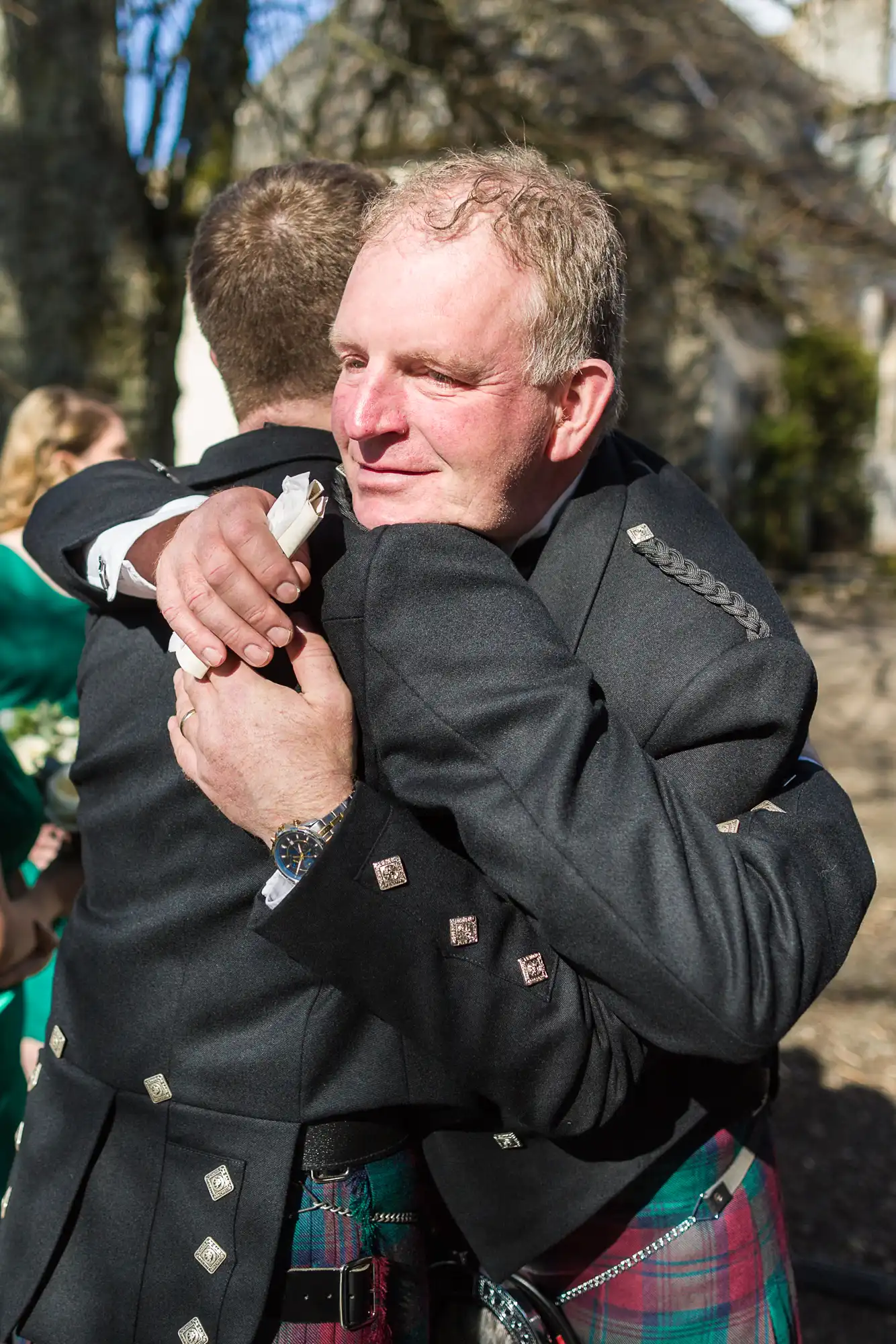 Two men wearing traditional Scottish kilts and dark jackets embrace warmly outdoors. One man’s face is visible, smiling with closed eyes, while the other’s back is to the camera. Sunlight highlights the joyful moment, with blurred trees in the background.