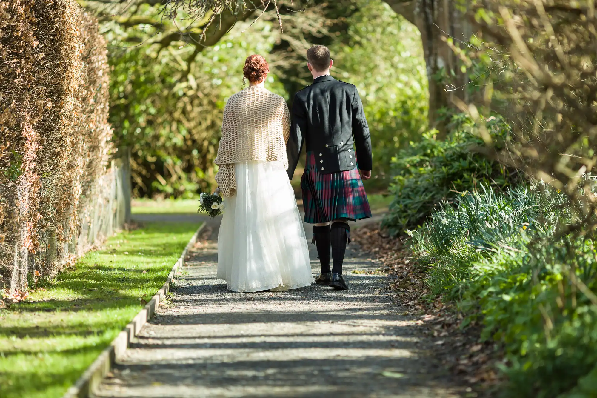 A couple walks hand in hand down a sunlit garden path. The woman wears a white dress and shawl, holding a bouquet, whilst the man wears a black jacket and tartan kilt. Trees, hedges, and greenery line the path on a bright day.