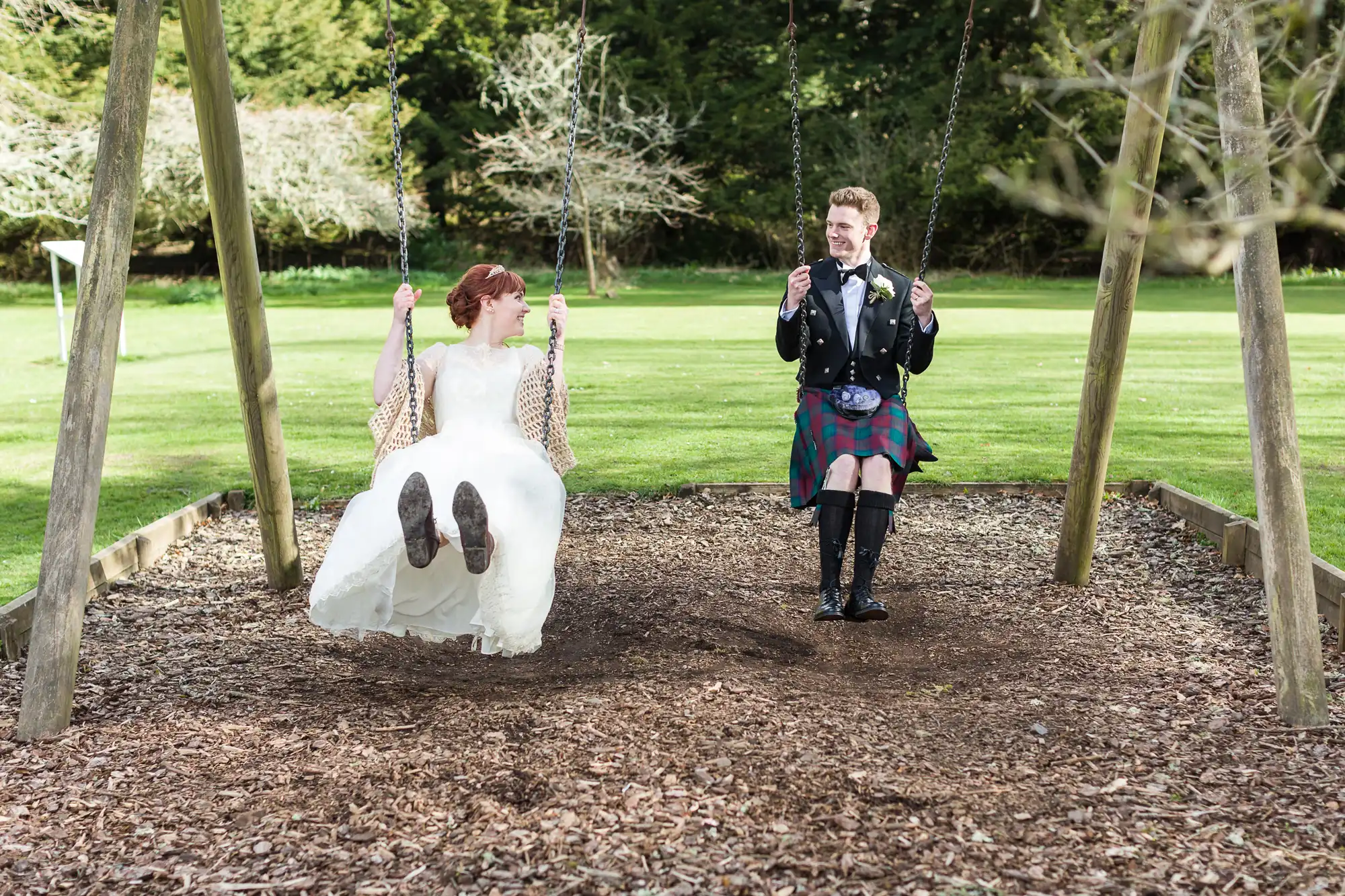 A bride in a white dress and a groom in a black jacket and tartan kilt sit on adjacent swings, smiling at each other, in a playground surrounded by grass and trees on a sunny day.