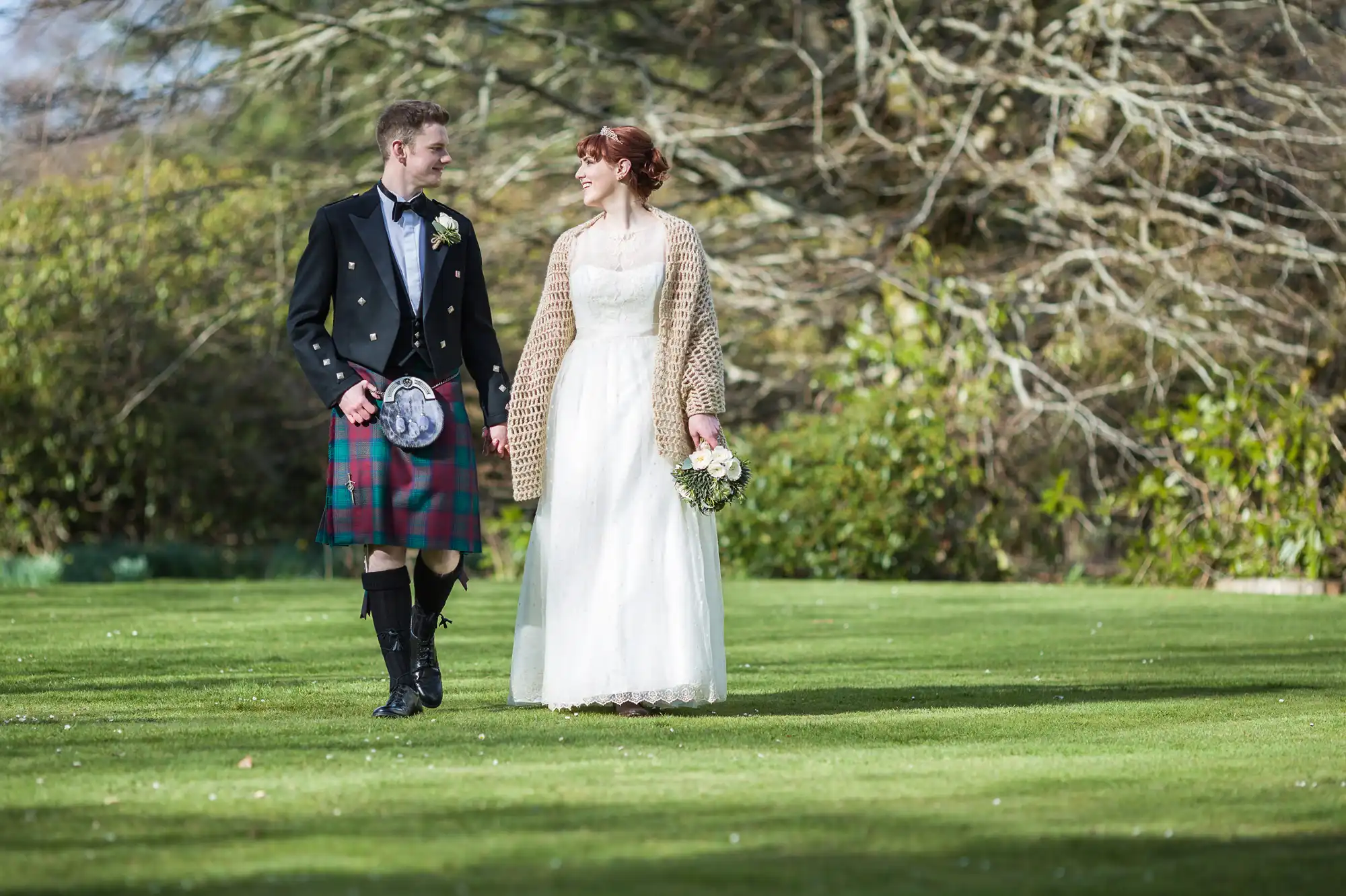 A smiling couple in wedding attire walk on grass. The groom wears a black jacket, kilt, sporran, and knee-high socks. The bride wears a white dress, cream shawl, and holds a bouquet. Trees with bare branches and greenery fill the background.