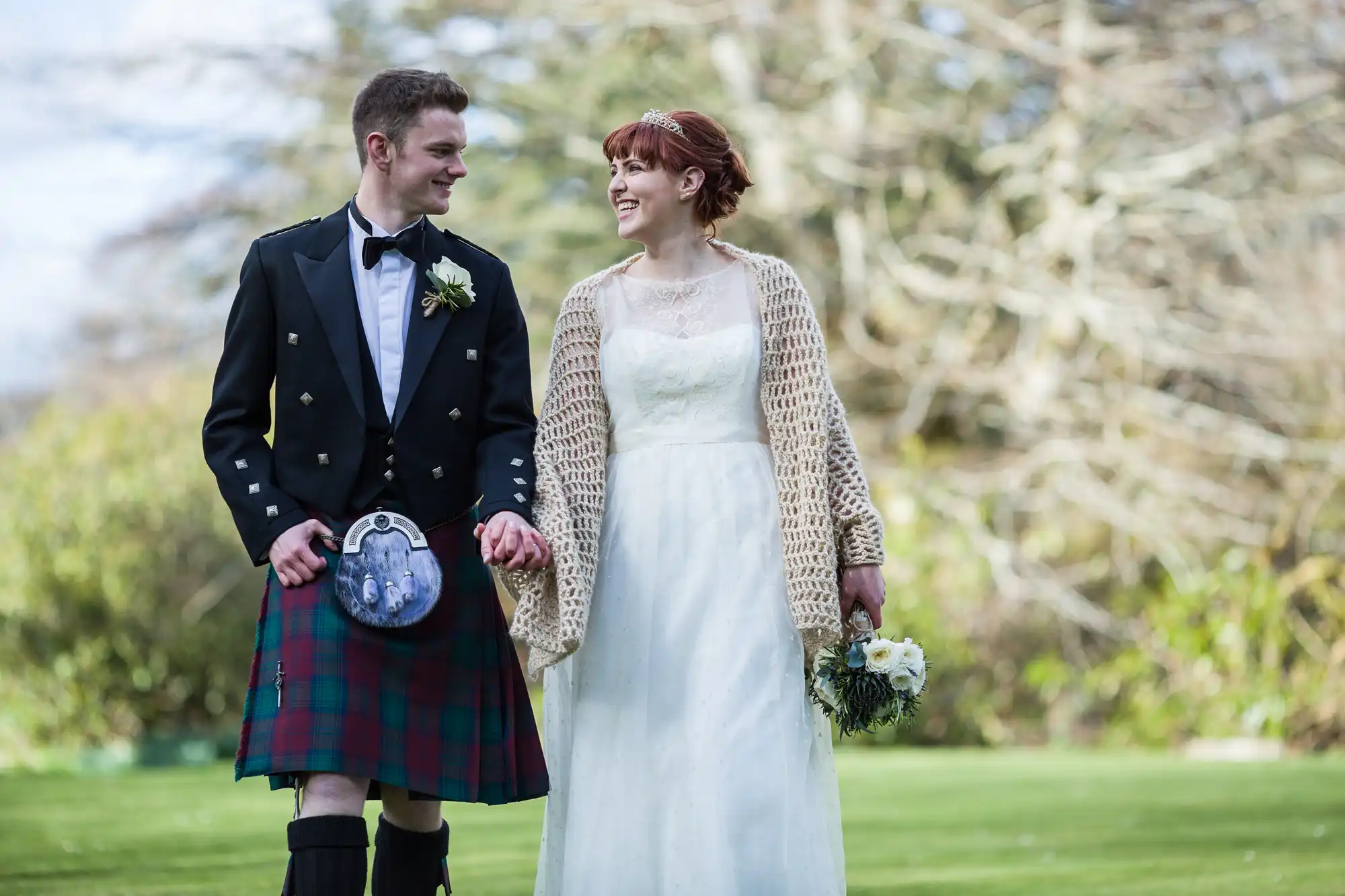 A smiling couple outdoors walk hand in hand. The man wears a black jacket, bow tie, and tartan kilt. The woman wears a white lace dress, beige knitted shawl, and holds a bouquet of flowers. Trees and grass fill the blurred background.