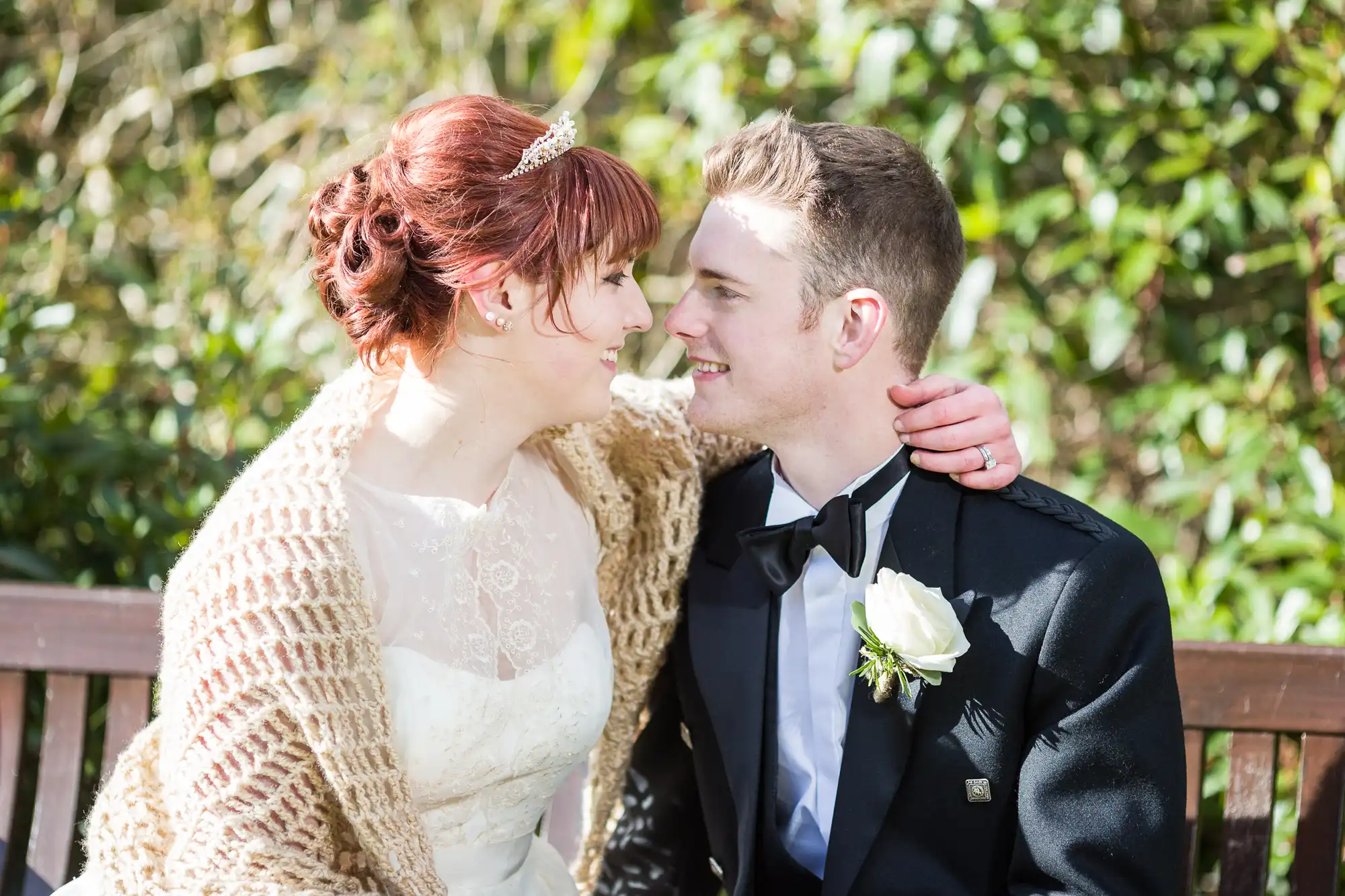 A bride and groom sit closely on a bench outdoors, smiling fondly at each other. The bride wears a white dress, tiara, and a shawl, while the groom wears a black suit with a white rose buttonhole. Sunlight highlights their happy faces with greenery in the background.