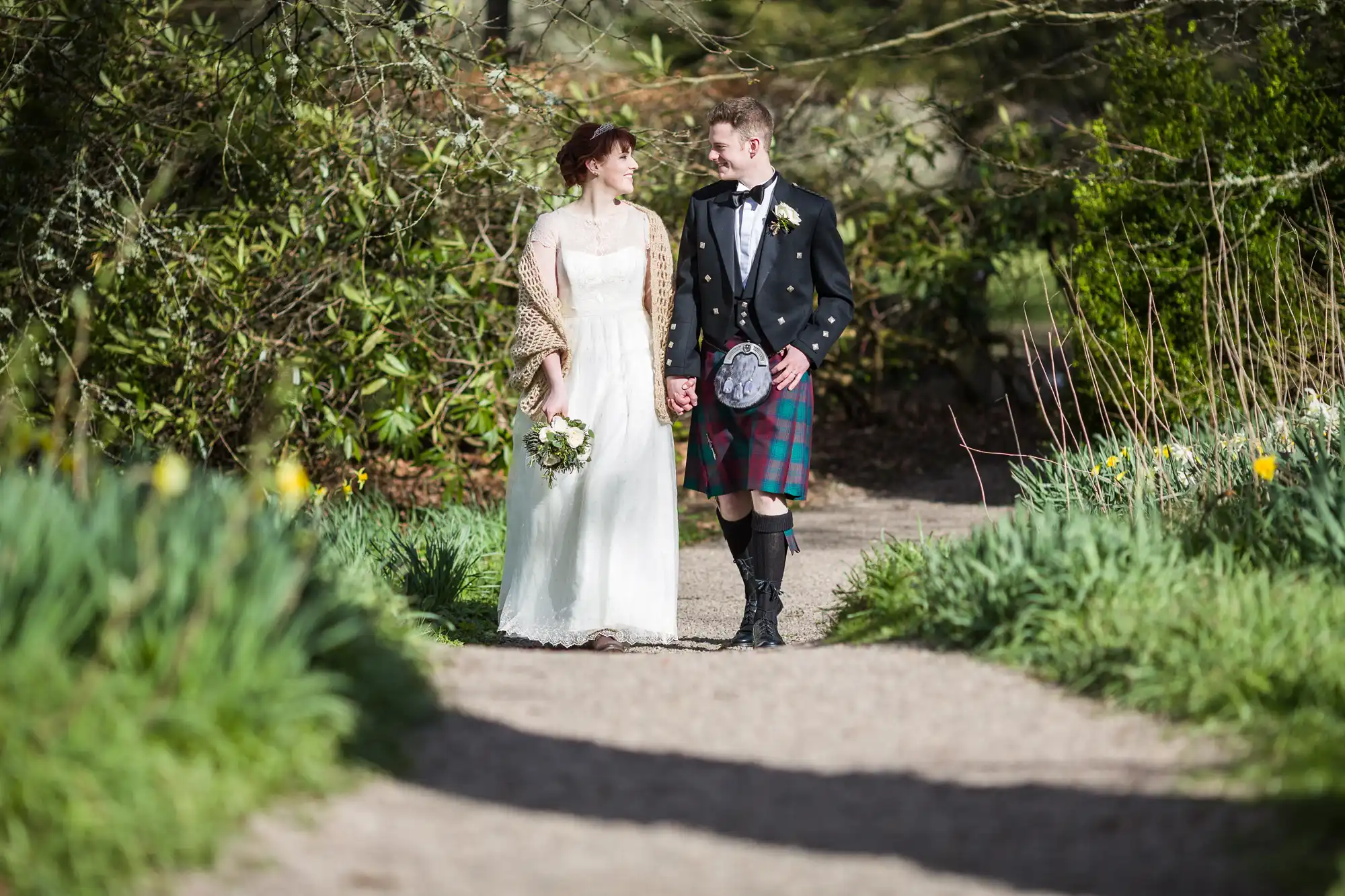 A bride in a white dress and shawl holds a bouquet and walks hand-in-hand with a groom in a black jacket and tartan kilt. They smile at each other on a sunlit garden path, surrounded by greenery and yellow flowers.