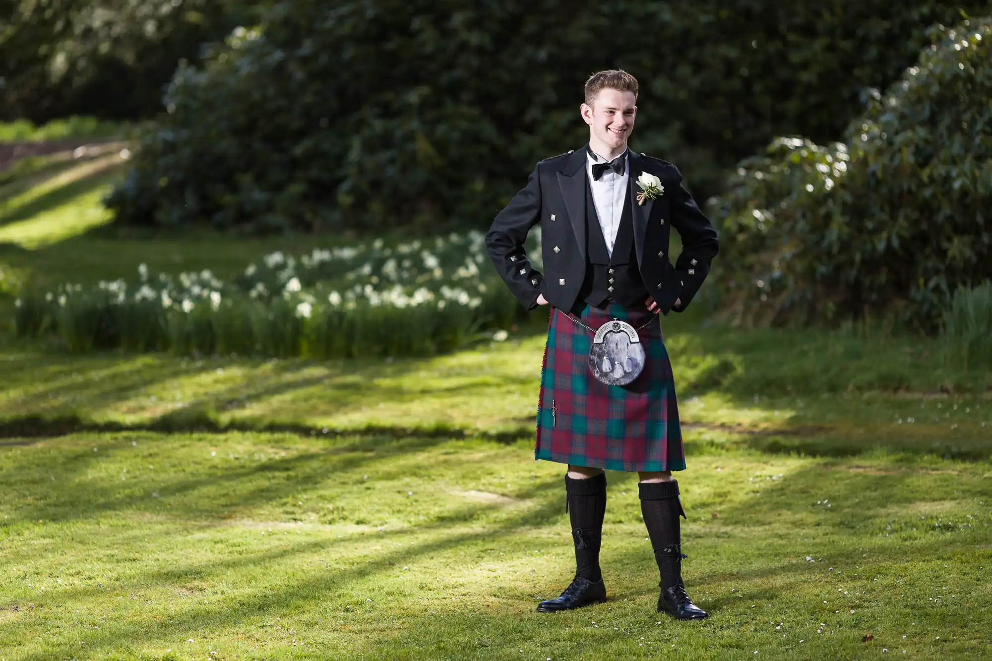 A young man stands on a sunlit lawn wearing traditional Scottish attire: a black jacket, white shirt with bow tie, red and green tartan kilt, sporran, kilt hose, and black shoes. He smiles with hands on hips. Trees and flowers are in the background.