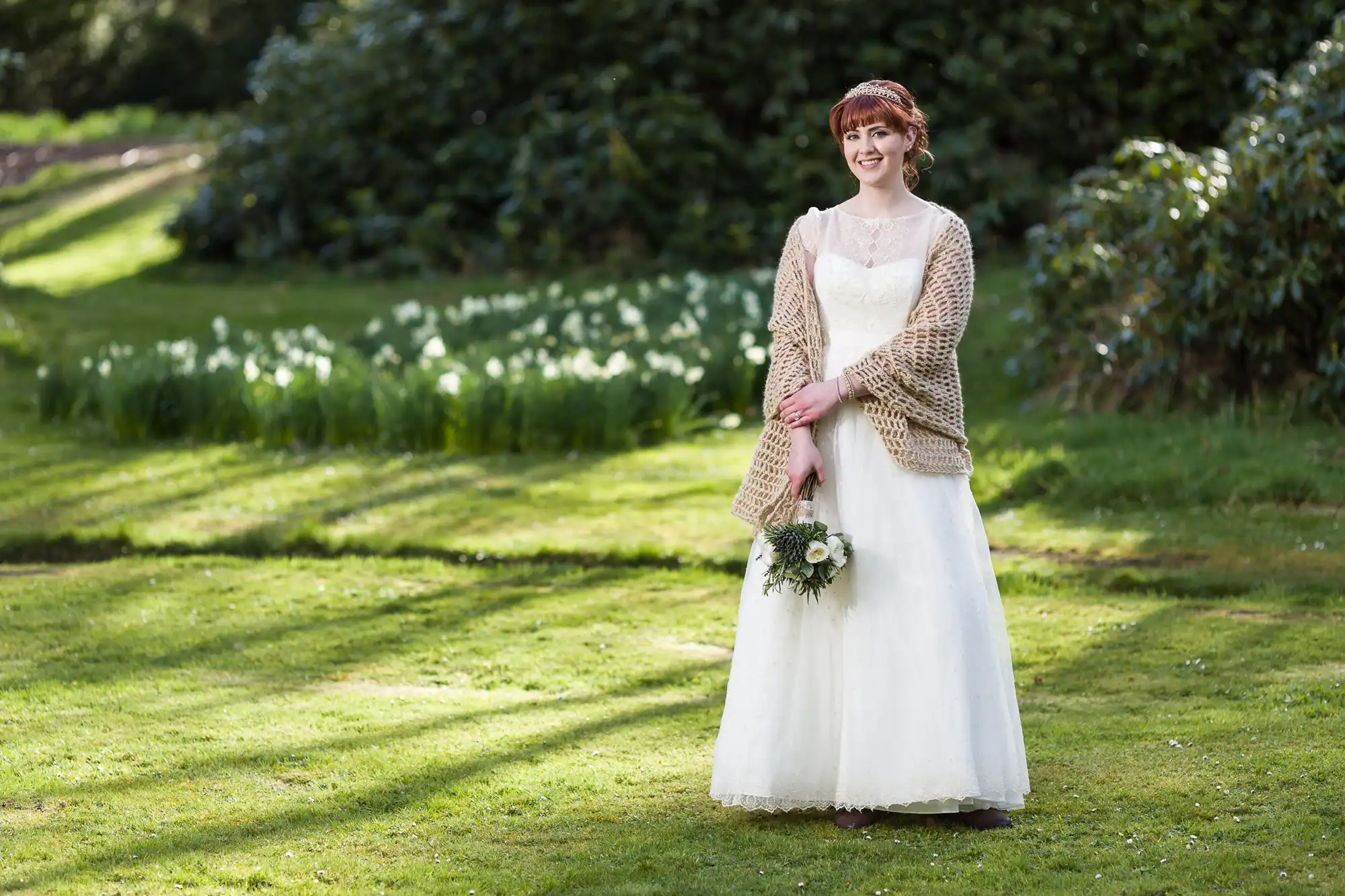 A smiling bride in a white gown stands on a sunlit lawn, holding a small bouquet. She wears a beige shawl over her shoulders. Behind her is a garden with green grass, blooming white flowers, and dense shrubs in the background.