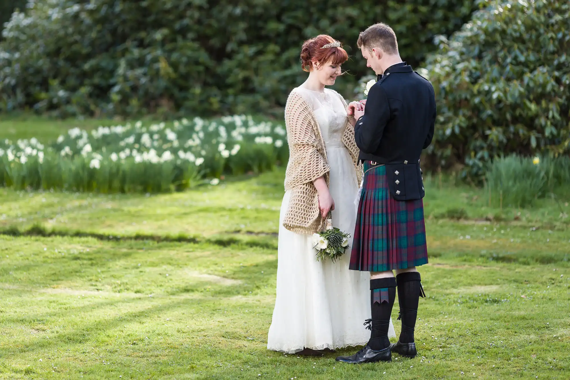 A bride in a white dress and tan shawl holding a bouquet stands facing a groom in a dark jacket and tartan kilt on green grass, with trees and white flowers in the background. Both are smiling and looking at each other closely.