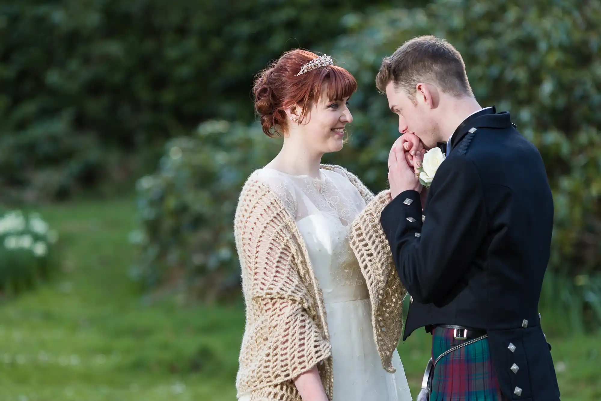 A bride in a white dress and light shawl smiles as a groom in a dark jacket and tartan kilt gently kisses her hand. They stand outdoors on green grass, with blurred trees and plants in the background, sharing an affectionate moment.