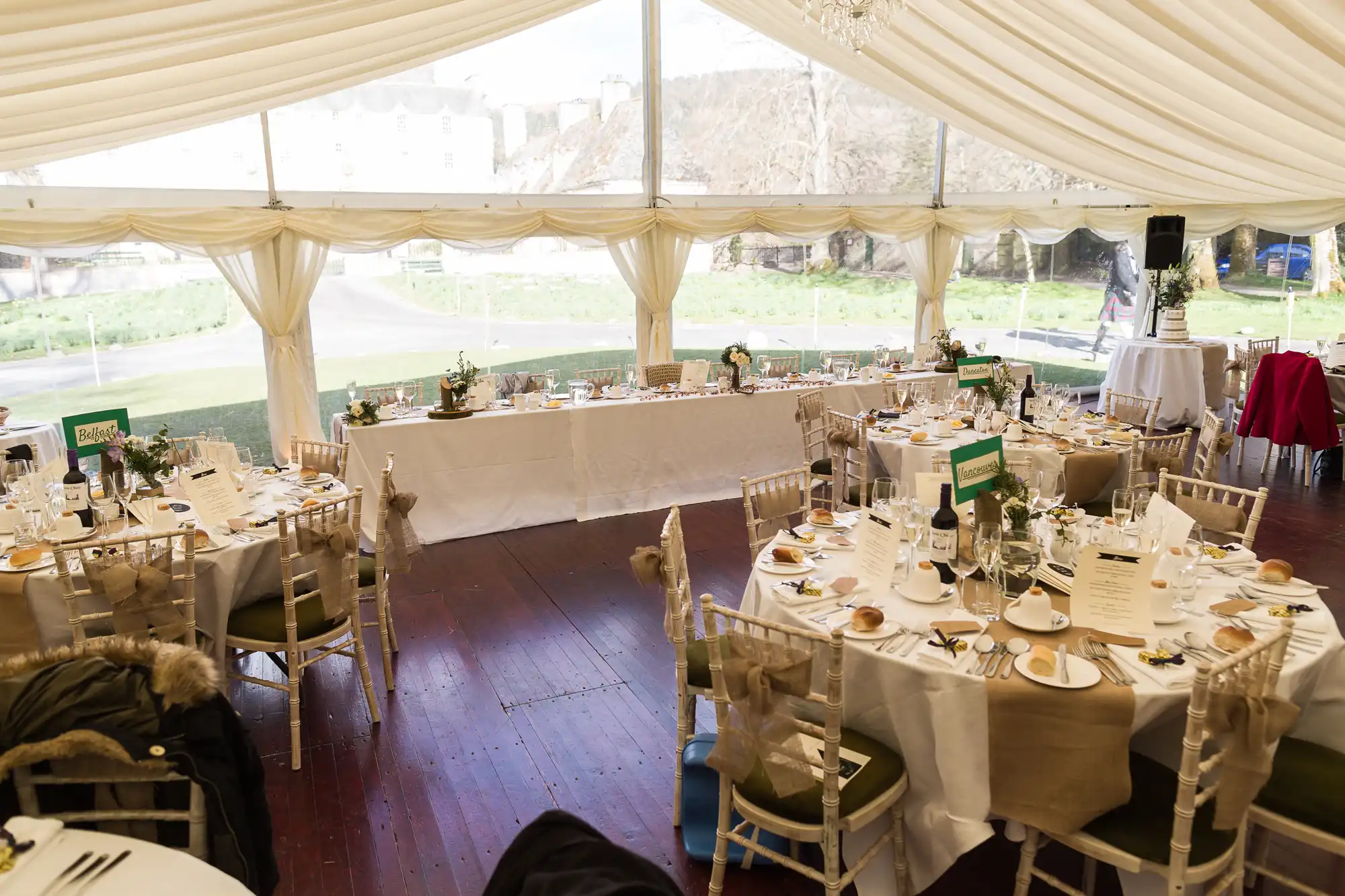 A bright wedding or event reception inside a large white marquee with round tables set for dining. Each table has neatly arranged plates, glasses, bread rolls, and centrepieces. A long top table is at the front, and daylight streams through clear marquee panels.