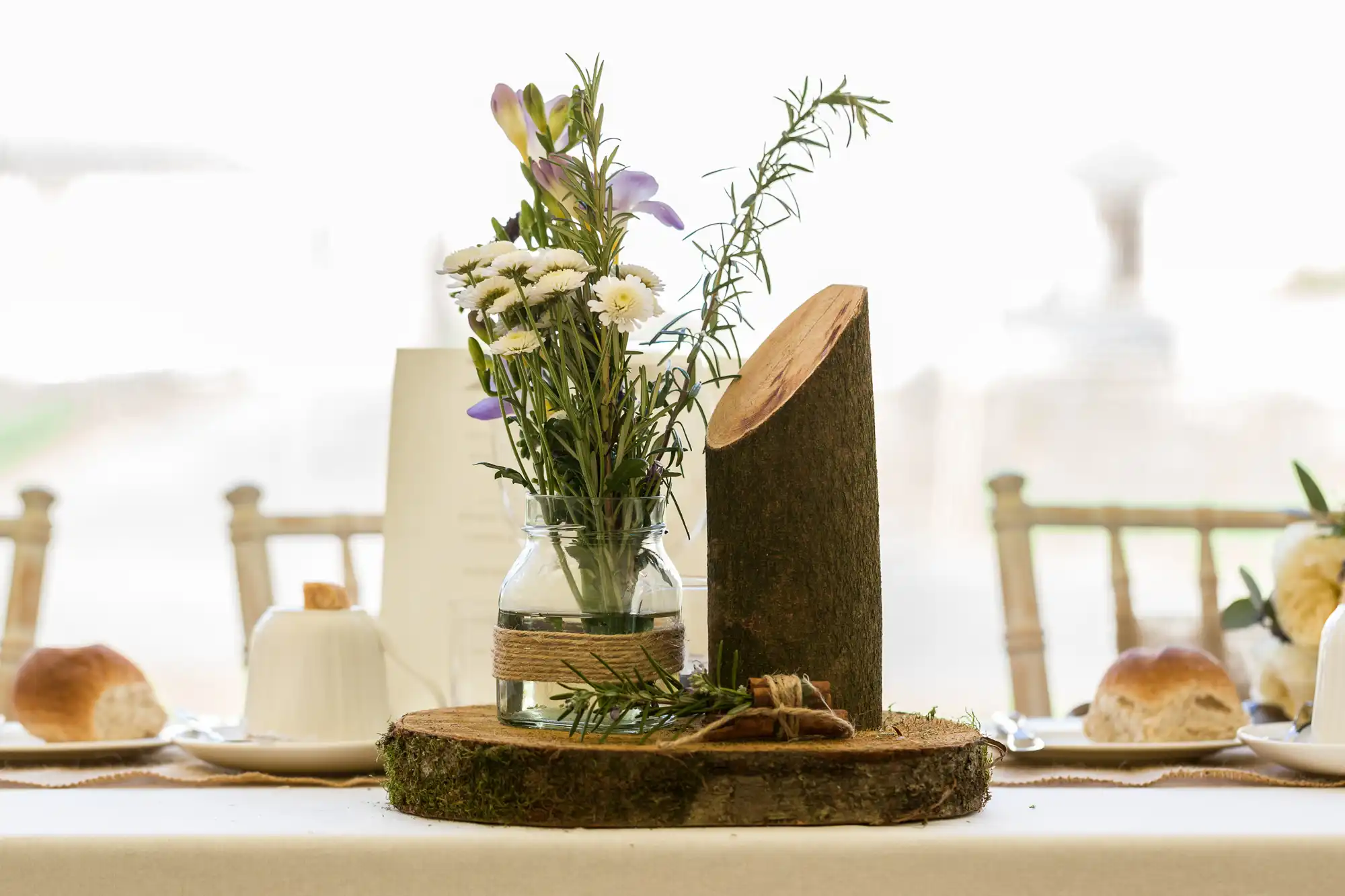 A rustic table centrepiece features a glass jar of white daisies and purple flowers, wrapped with twine, next to vertical and horizontal wood slices. The scene includes white plates, teacups, and bread rolls on a white tablecloth.