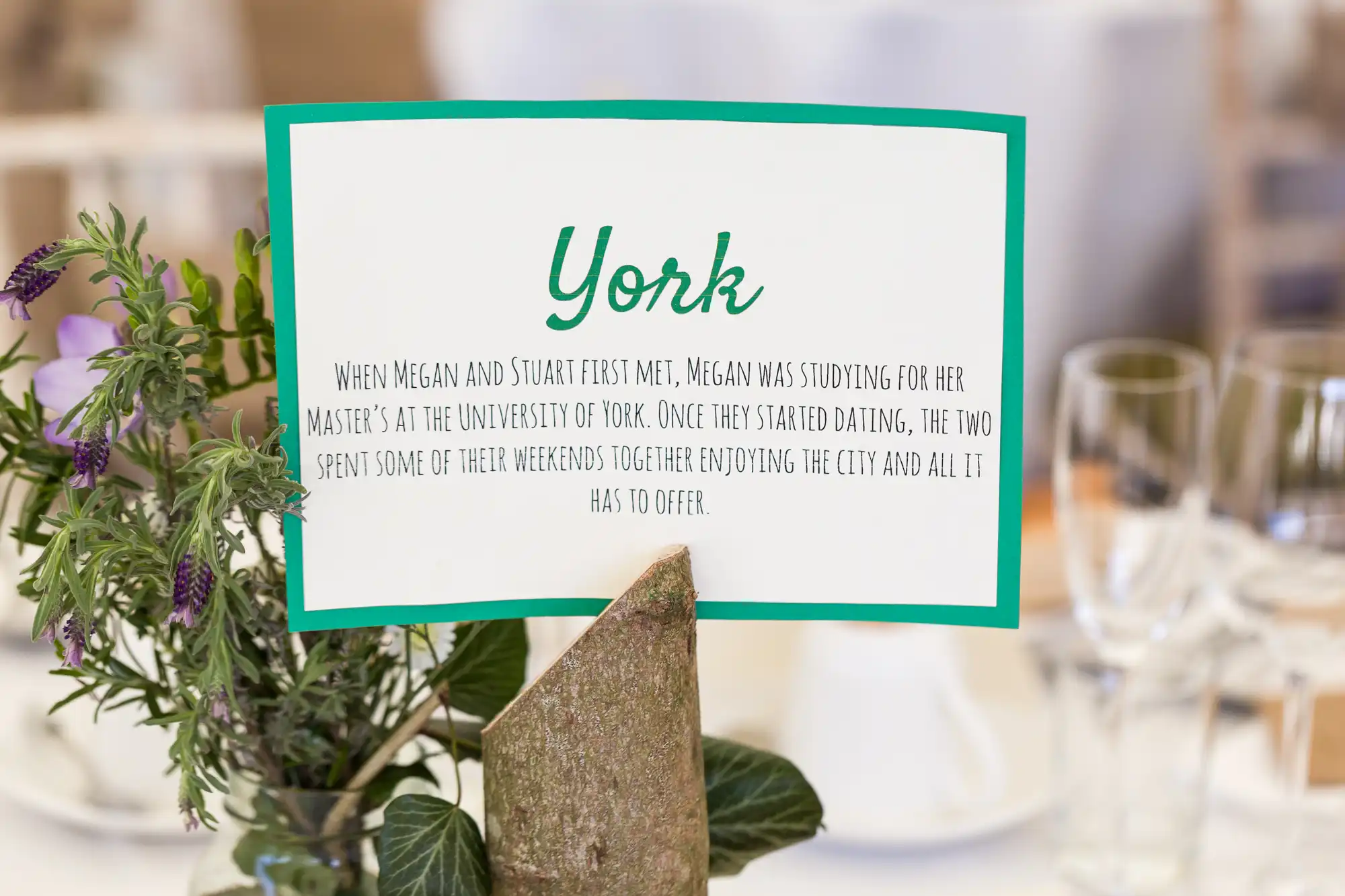 A table sign titled "York" describes Megan and Stuart spending weekends together in York while Megan studied for her master’s. The sign is propped among green, purple, and white flowers. Glasses and plates are blurred in the background.