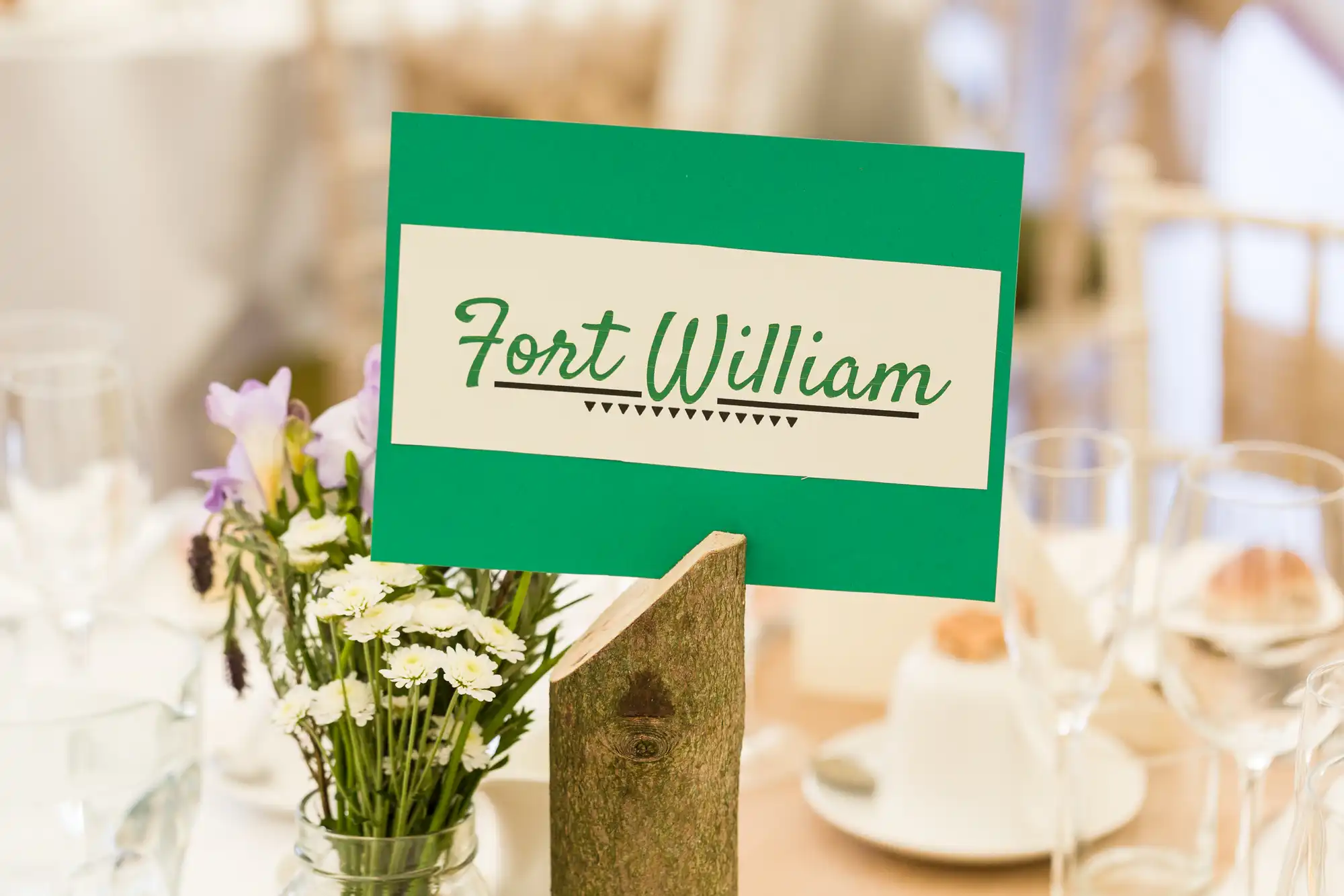 A green and white sign reading "Fort William" is displayed on a wooden stand. The background features a table set with glassware, plates, and a small bouquet of white and purple flowers in a vase, creating a festive ambience.
