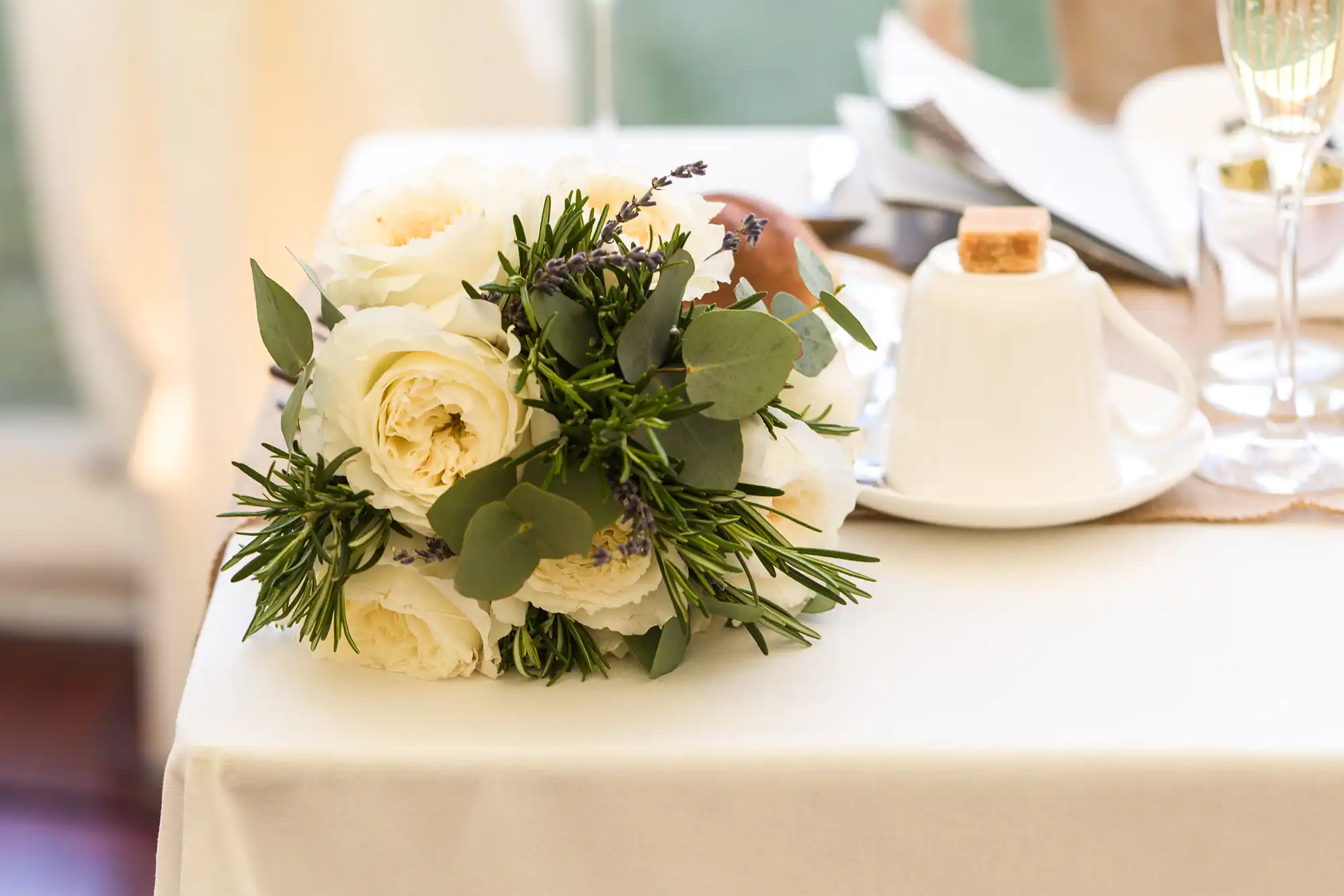 A small posy of white roses, lavender, and green foliage rests on a white tablecloth beside a white cup with a sugar lump on top, silver cutlery, a champagne glass, and a folded serviette on a set dining table.