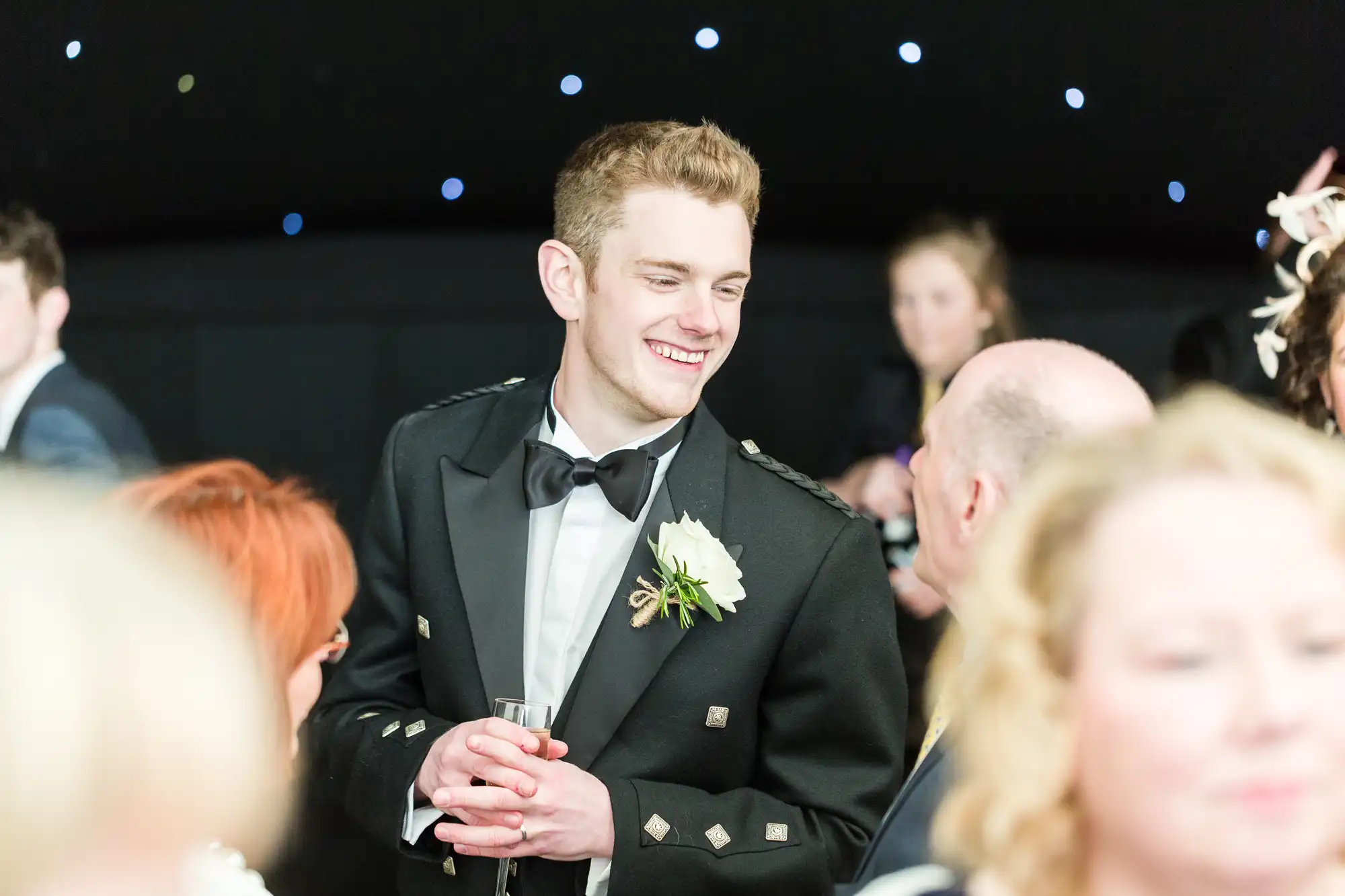 A young man in a black dinner jacket with a bow tie and white flower buttonhole smiles and holds a drink whilst chatting with others at a formal event. The background is softly lit with small, star-like lights.