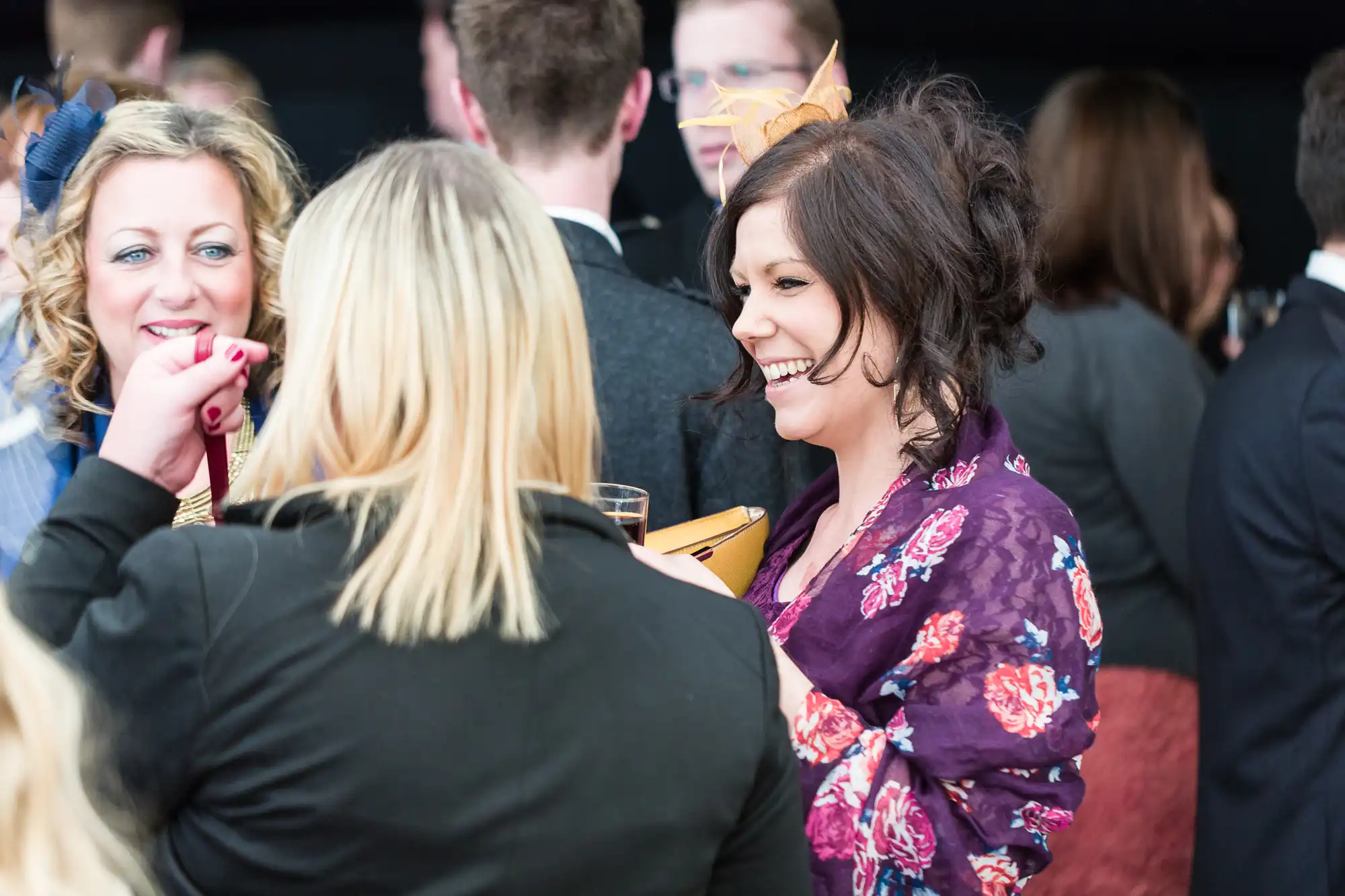 A group of women in colourful, dressy attire chat and laugh at a crowded indoor event. One woman, smiling, wears a purple floral shawl and holds a yellow clutch bag. People in suits and dresses mingle in the background, facing away.