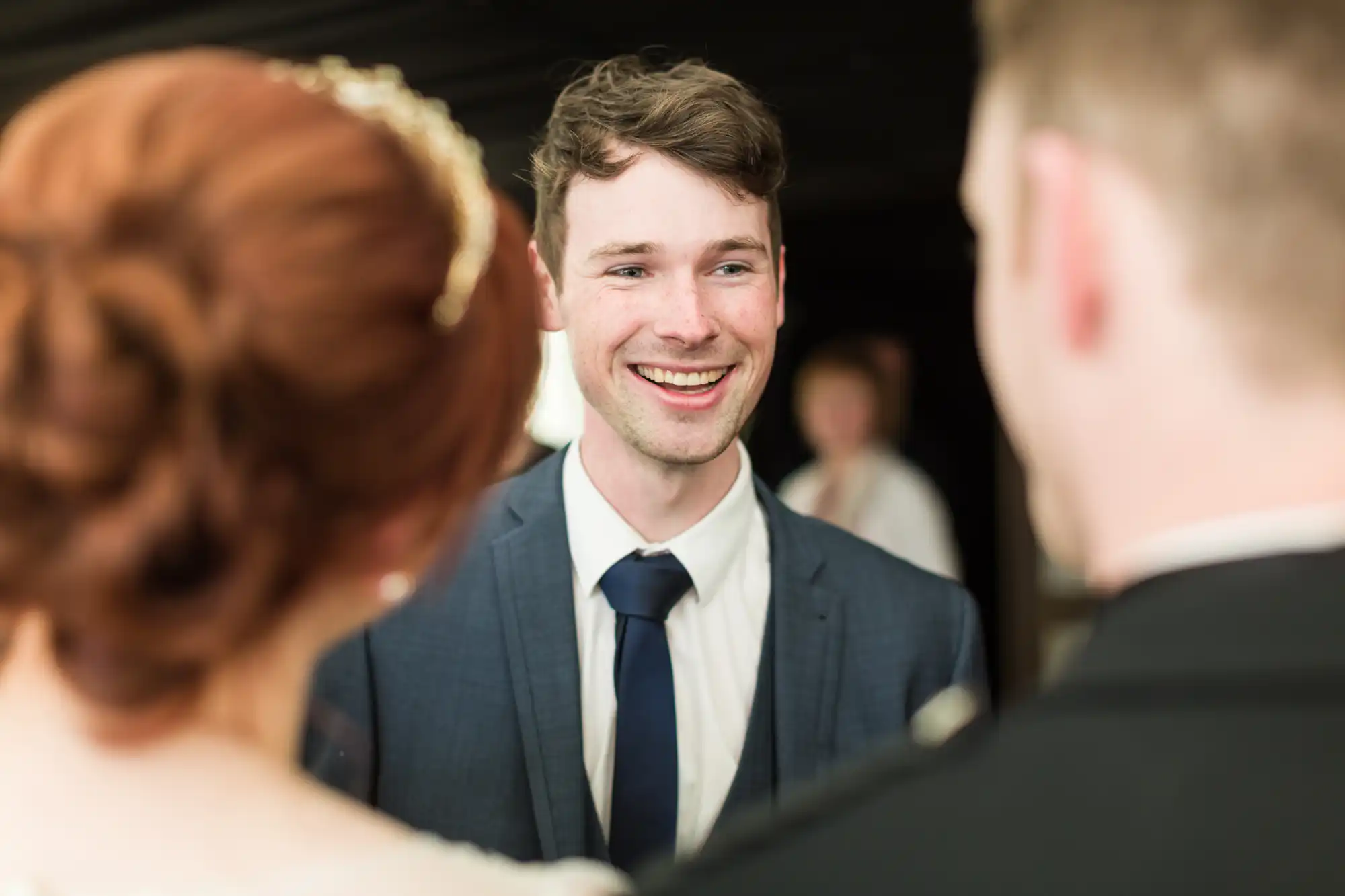 A young man in a blue suit and tie smiles warmly at two people in the foreground, who are slightly out of focus. The man has light skin and brown hair. The background is dark, with another blurred person visible.