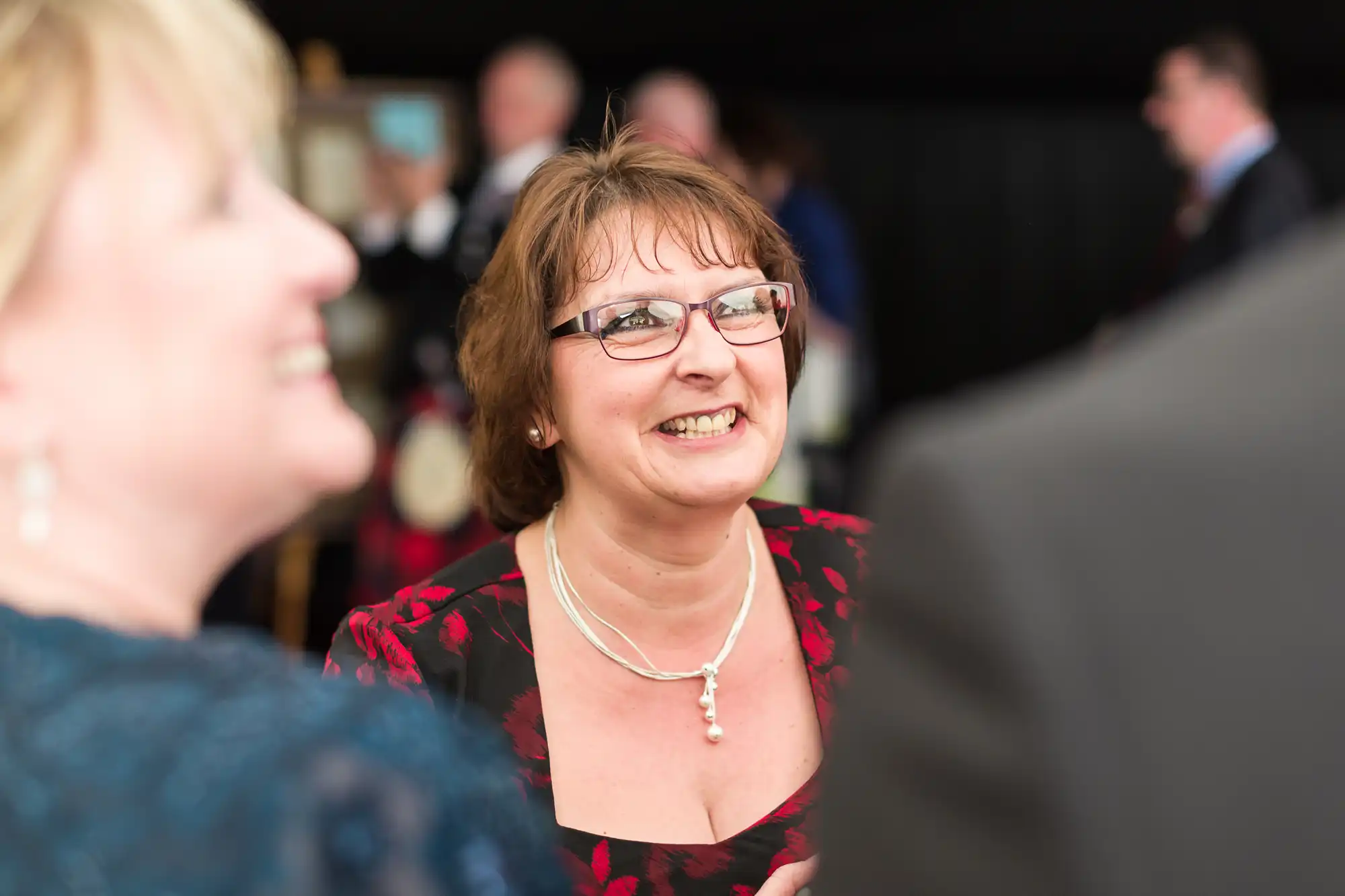 A woman with short brown hair, glasses, and a red-and-black dress smiles whilst talking to others at a social event. She wears a pearl necklace. The background is blurred, with other people visible but out of focus.