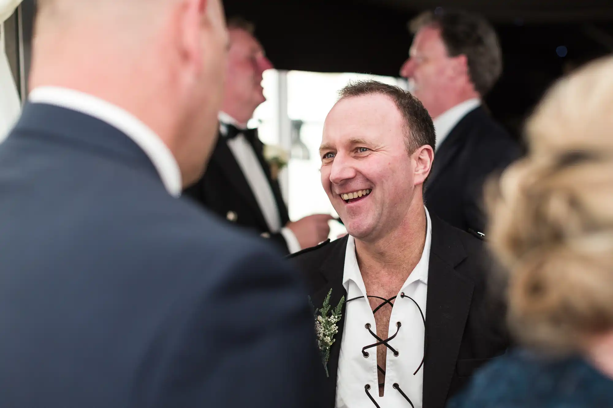 A man in a black jacket and white shirt with a lace-up collar laughs whilst conversing with others at a formal event. Two blurred people are in the foreground, and another man in a dinner jacket is visible in the background. The mood is cheerful and sociable.