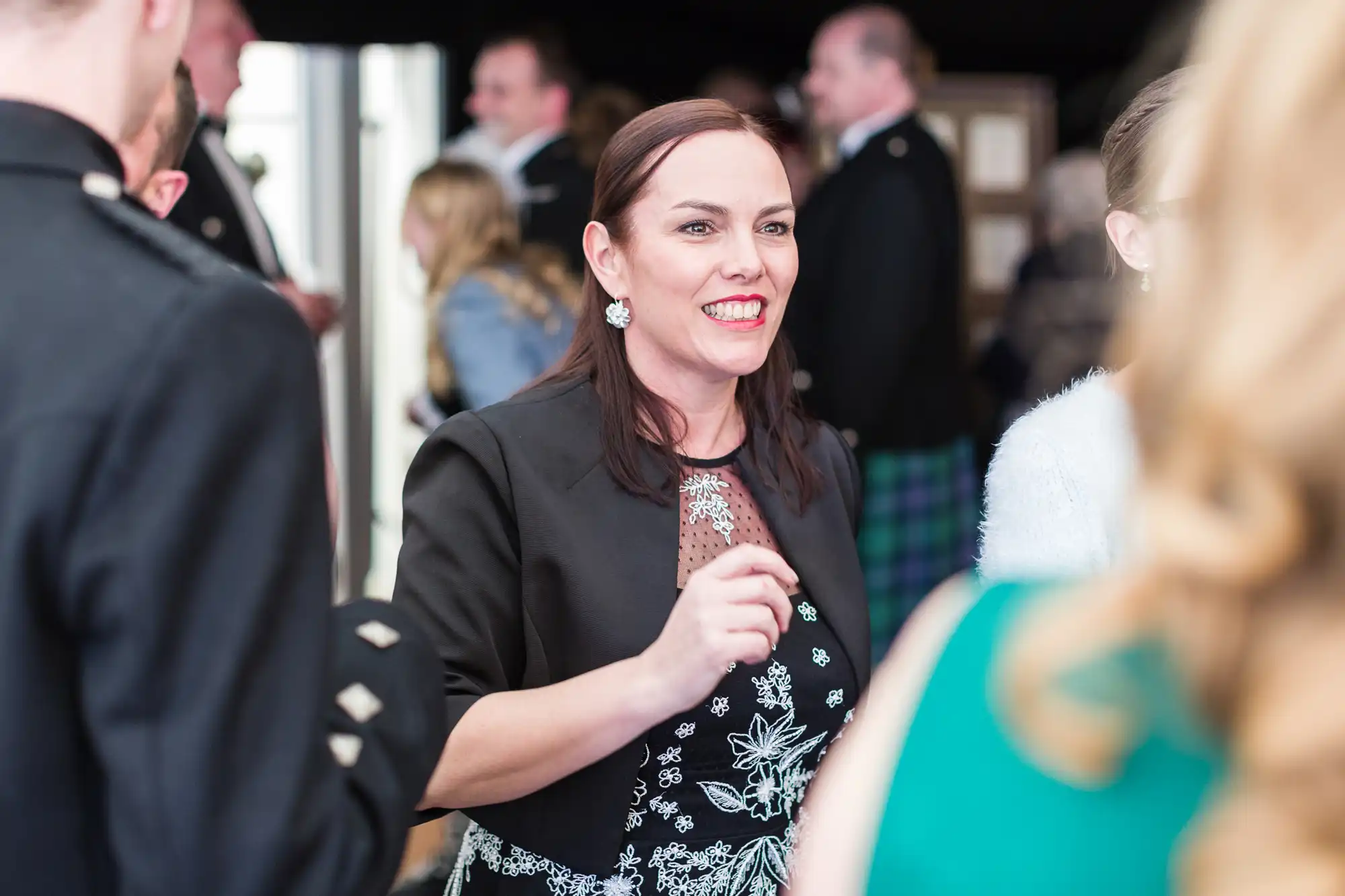 A woman with brown hair wearing a black jacket over a patterned dress smiles and gestures whilst talking to people at a formal event. Others in suits and dresses are visible around her, with blurred figures and a festive, lively atmosphere in the background.