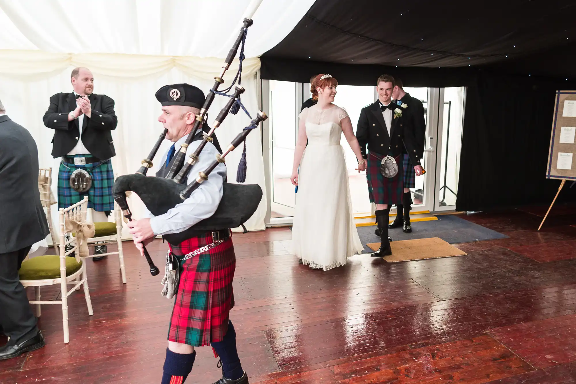 A bagpiper in traditional Scottish attire leads a bride in a white dress and groom in a kilt into a marquee with wooden floors. Other guests in kilts and formal wear are clapping. The scene suggests a Scottish wedding celebration.