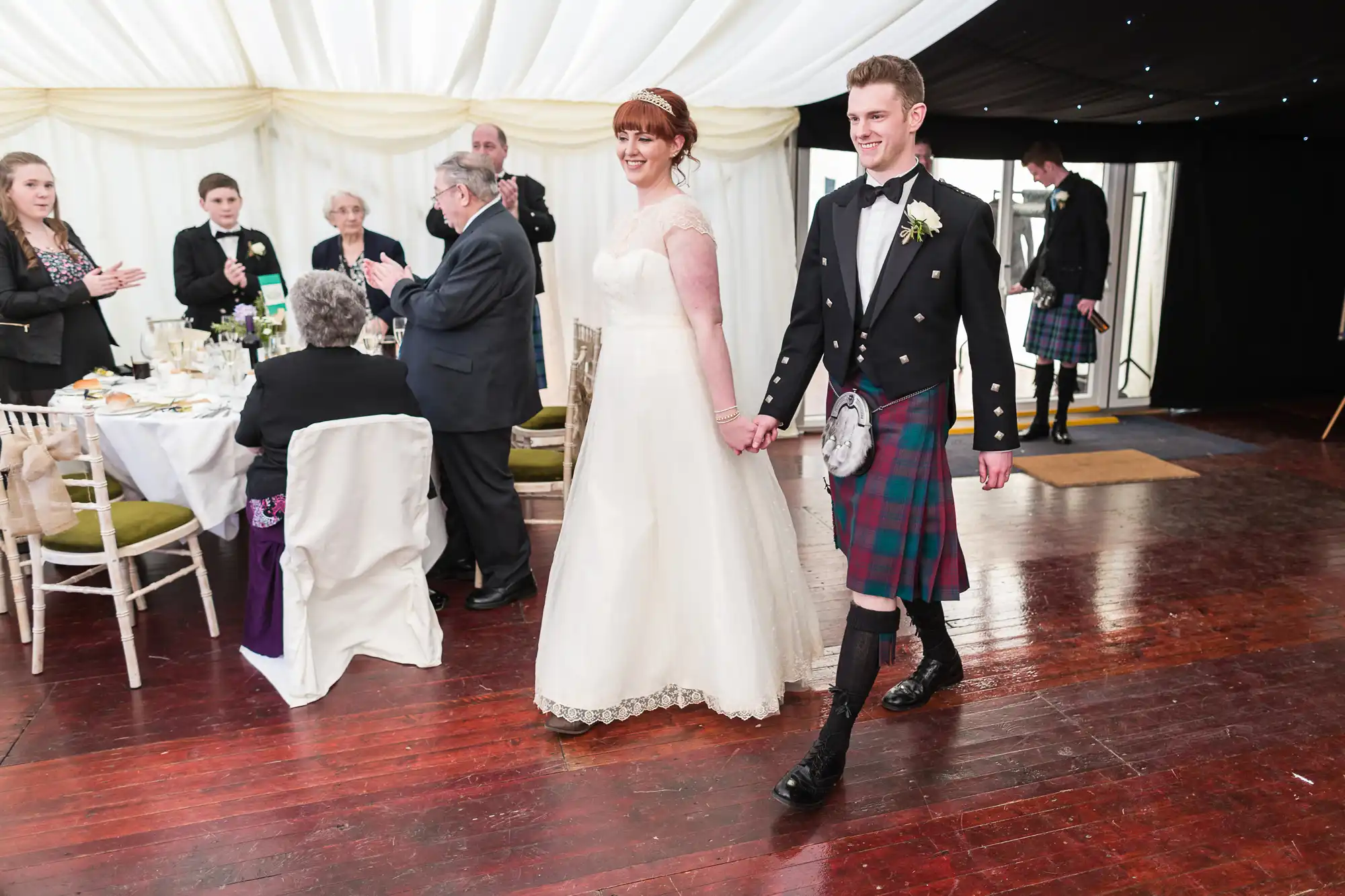 A smiling bride in a white gown and a groom in a kilt and formal Scottish attire walk hand-in-hand in a marquee, as seated and standing guests applaud. The wooden floor contrasts with the white draped ceiling and banquet-style tables in the background.