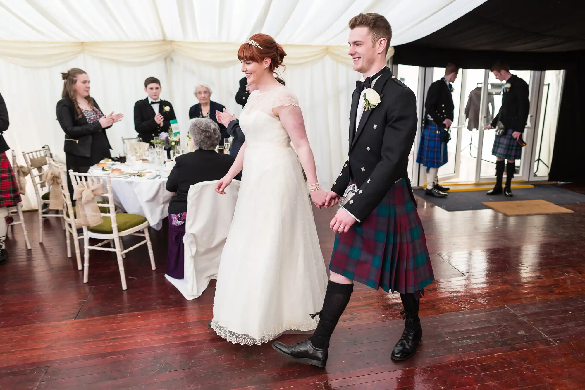 A bride in a white gown and a groom in a black jacket and tartan kilt walk hand in hand across a wooden floor in a wedding reception marquee, with seated and standing guests in the background, some wearing kilts.