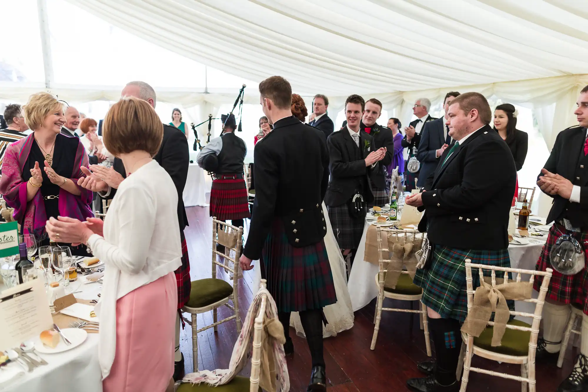 A group of people, many men wearing kilts and jackets, are standing and conversing around dining tables in a light-filled marquee. The tables are set for a formal meal, and the mood appears celebratory, likely at a wedding or special occasion.