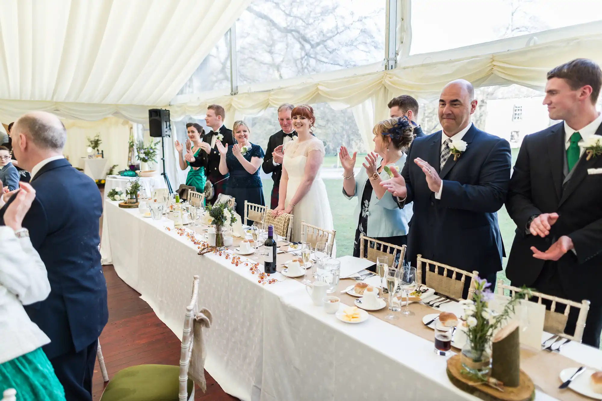 A wedding reception scene with guests in formal attire standing and applauding around a long, decorated table. A bride in a white dress and groom in a black dinner suit stand in the centre, smiling. Plates, glasses, and floral centrepieces are on the table.