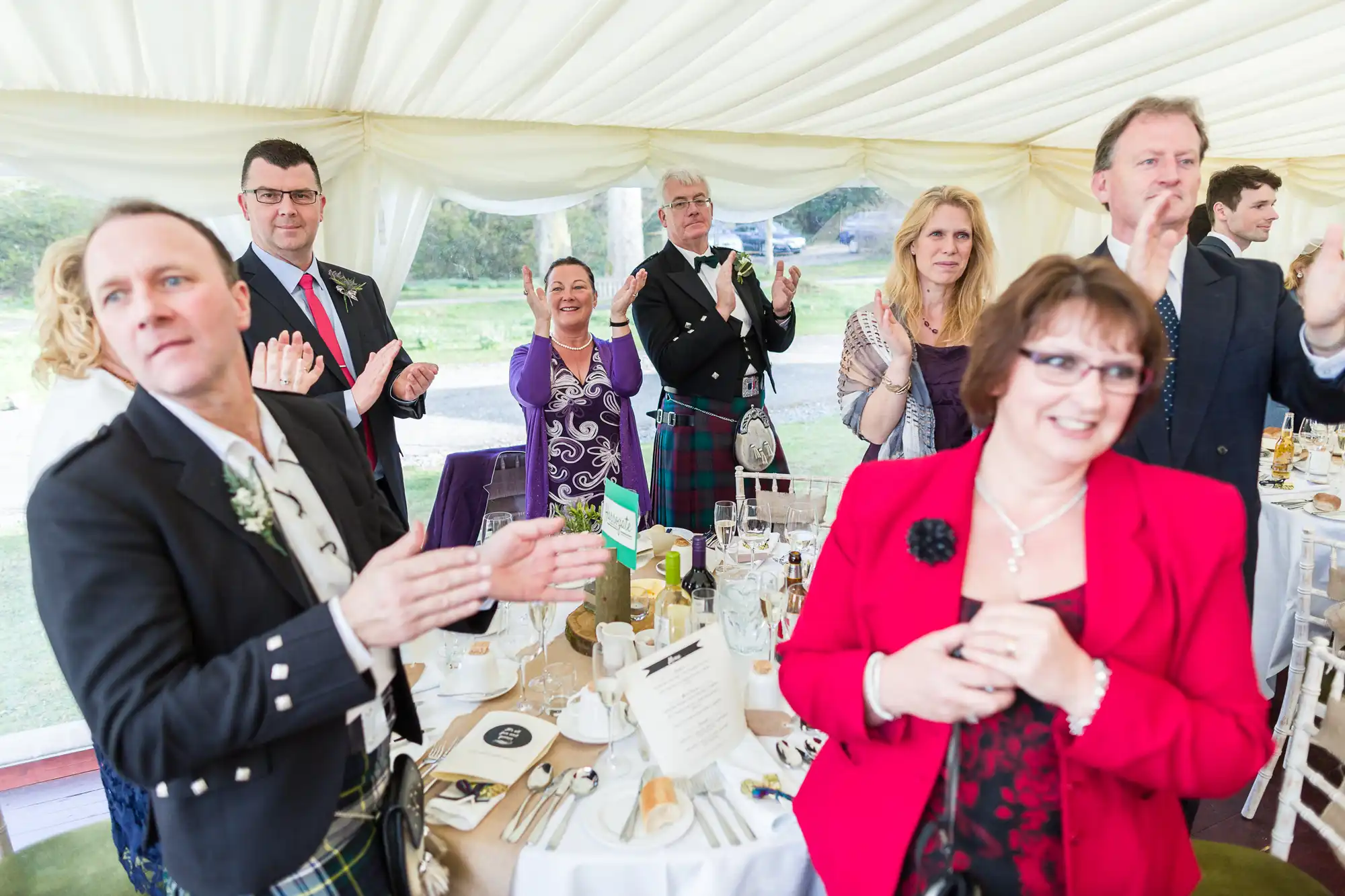 A group of well-dressed people stand around a decorated dining table under a white marquee, clapping and smiling. Some men wear kilts and jackets, and women wear dresses and blazers. Plates, menus, and drinks are on the table. The atmosphere is festive.