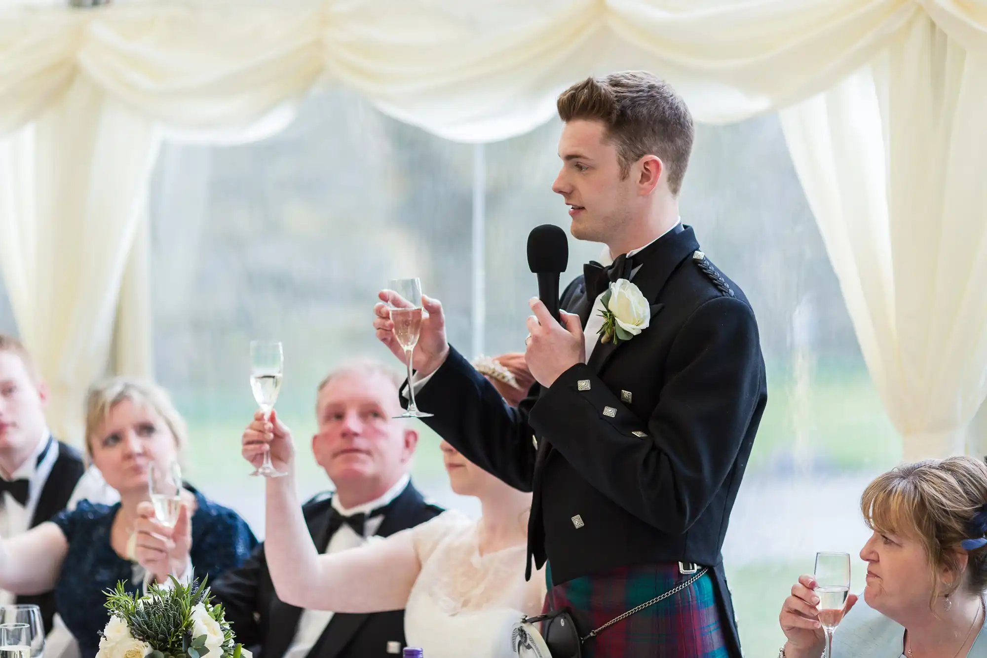 A man in a kilt with a white rose buttonhole stands holding a microphone and a champagne glass, giving a toast at a wedding reception. Seated guests in formal attire raise their glasses and look at him, with a decorated table and white draped background.