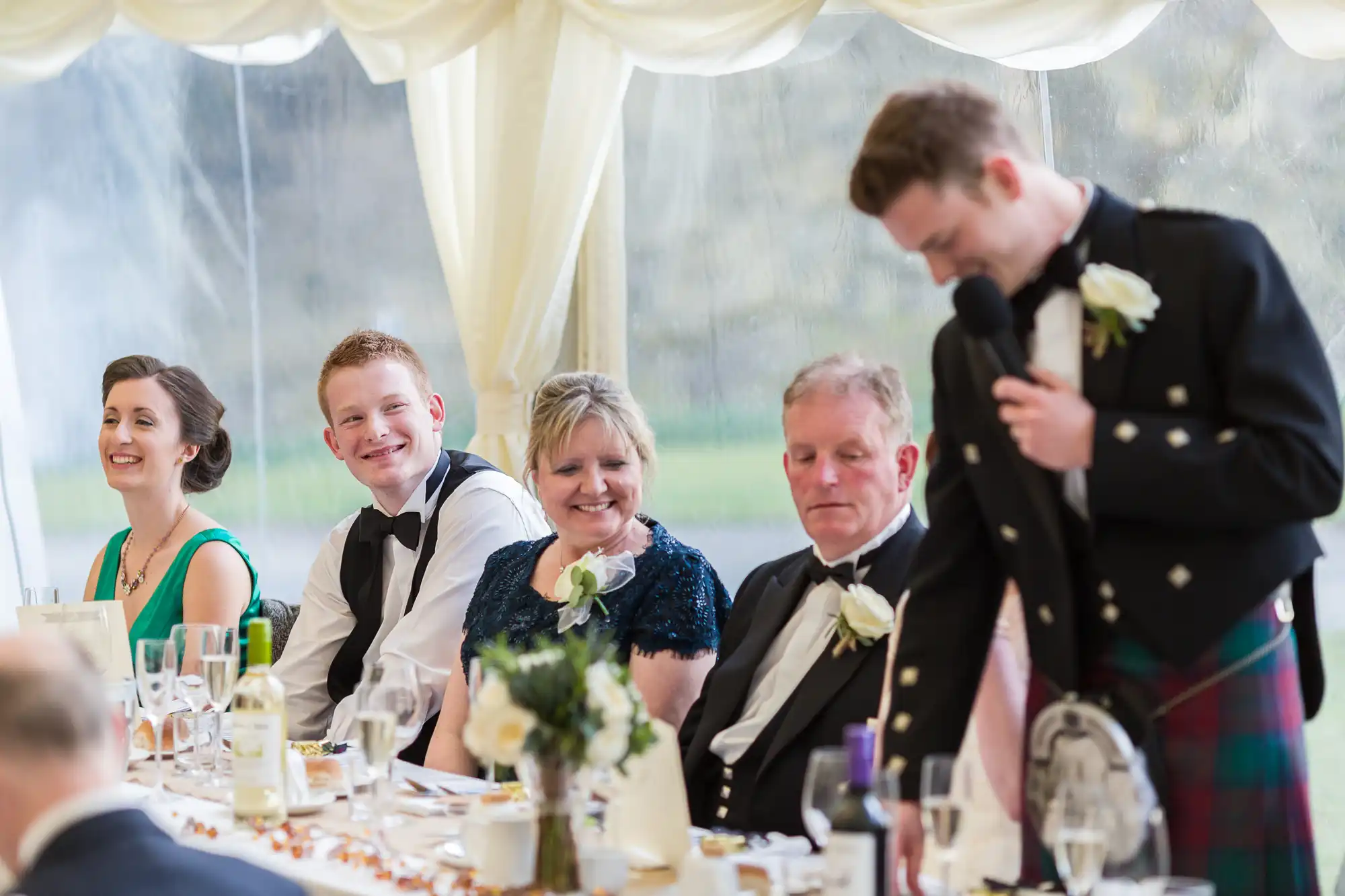 A group of wedding guests, dressed formally, sit at a decorated table inside a marquee. Four people are seated, smiling and looking at a standing man in a kilt holding a microphone, likely giving a speech. There are flowers, glasses, and bottles on the table.