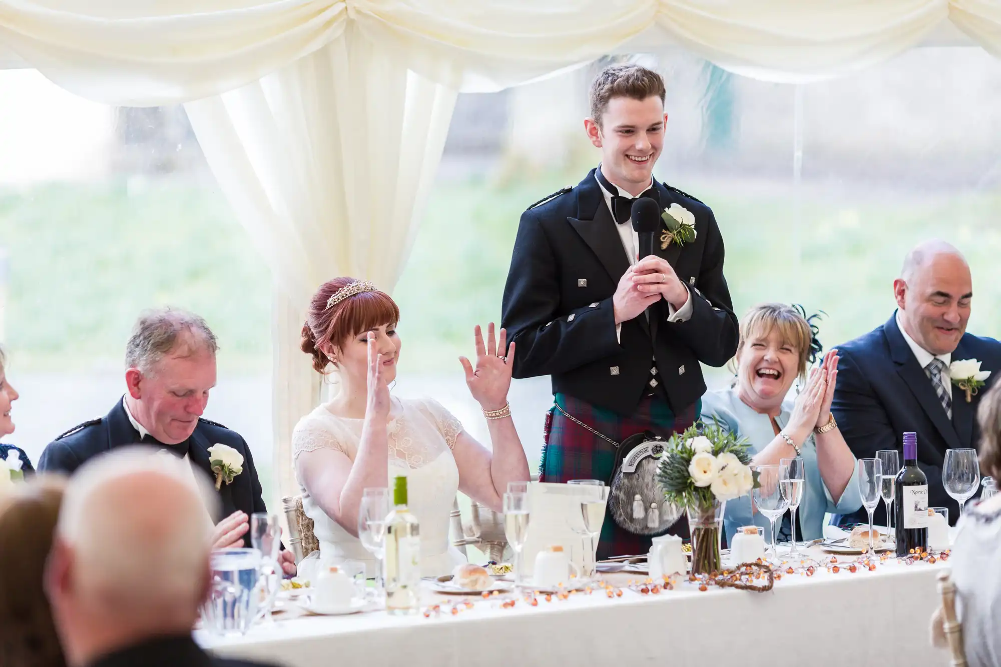 A young man in a kilt stands and speaks into a microphone at a wedding reception table. Guests, including a woman in a white dress and veil, are seated beside him, smiling and laughing. The table is decorated with flowers, glassware, and drinks.
